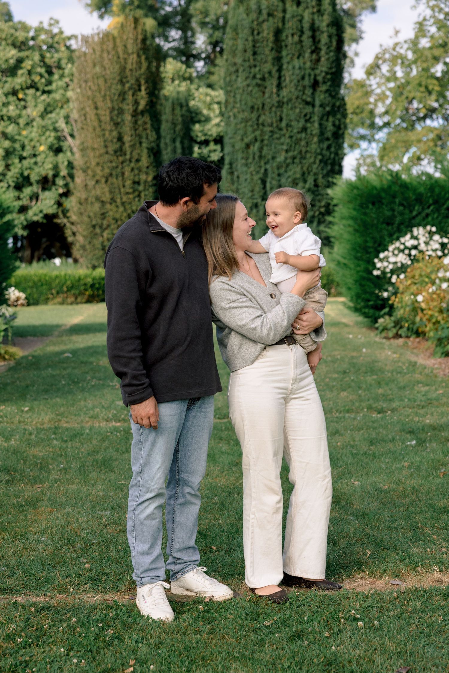 A happy young family with a baby boy poses in a lush green garden with tall cypress trees.