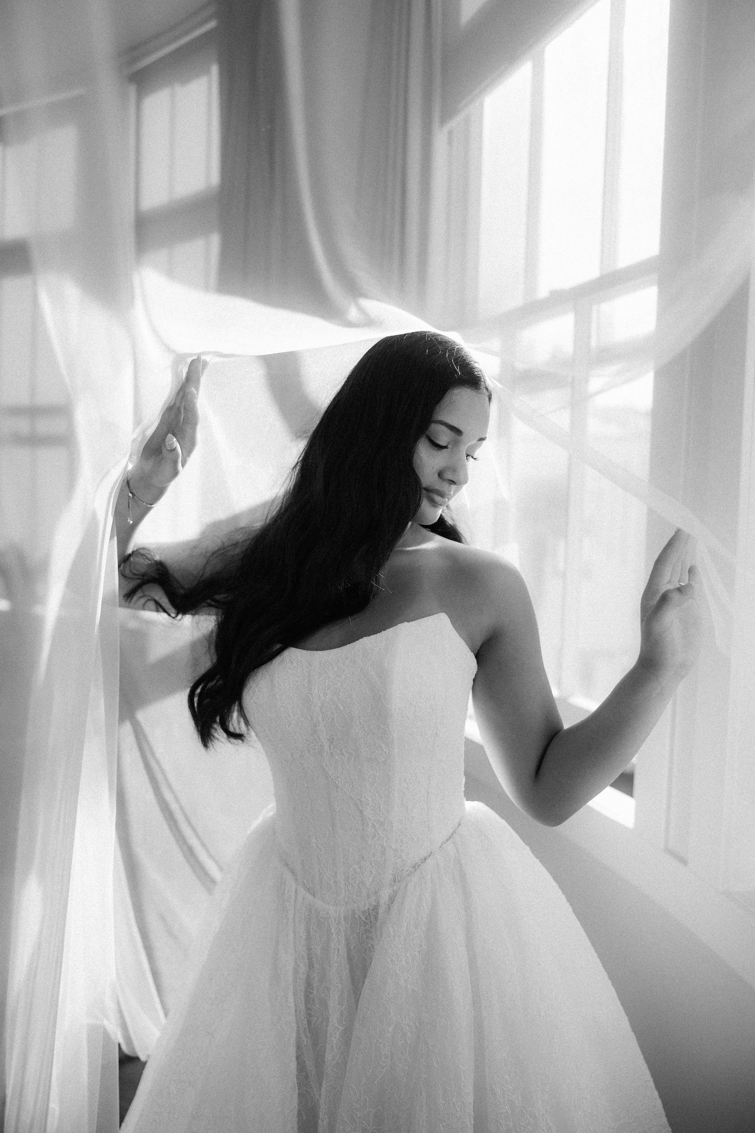 Black and white photo of a bride in a strapless gown holding flowing white curtains near a bright window.