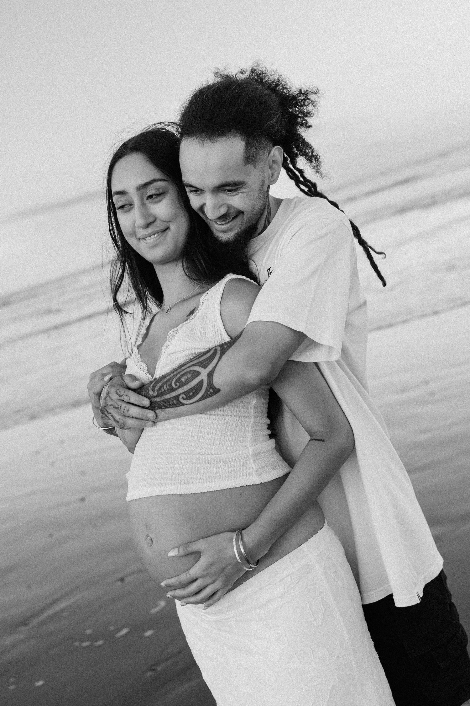 Black and white photo of smiling couple on beach, man with dreadlocks embracing pregnant partner.