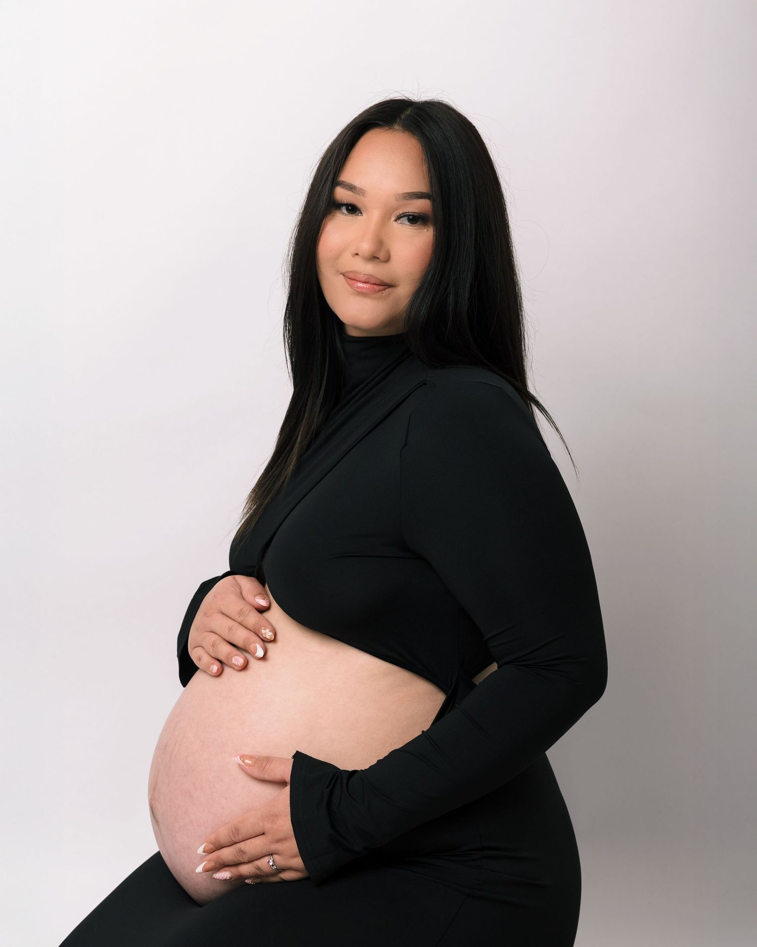 Pregnant woman in black turtleneck dress holding her bare baby bump in a studio portrait.