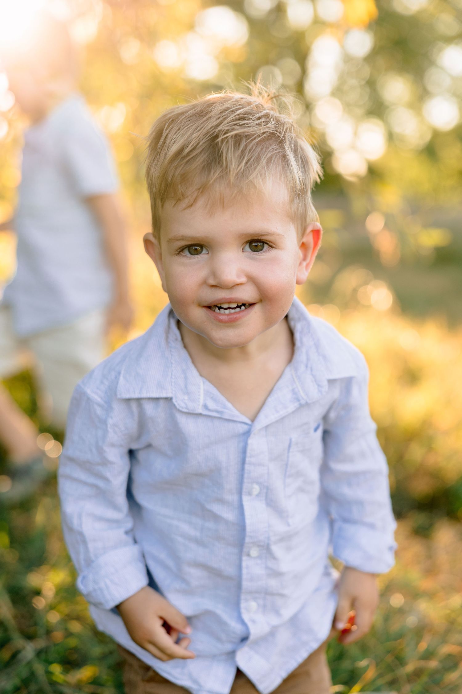 Smiling blonde toddler boy in light blue linen shirt standing outdoors in golden sunlight.