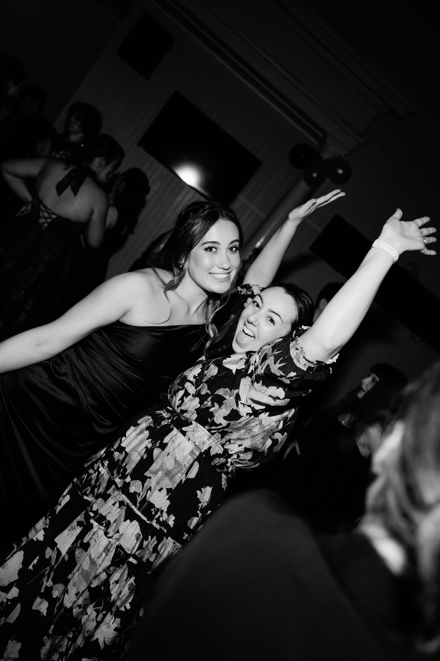 Two women dancing joyfully with arms raised at a formal event, captured in black and white photography.