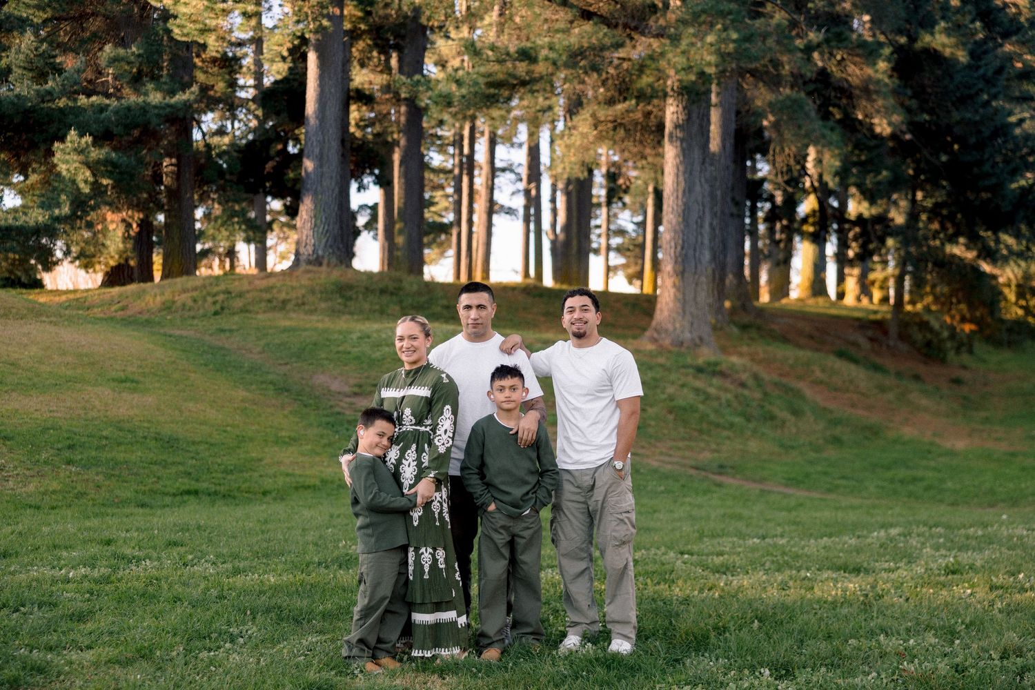 Family of five posing together on a green lawn surrounded by tall trees in a park during golden hour.