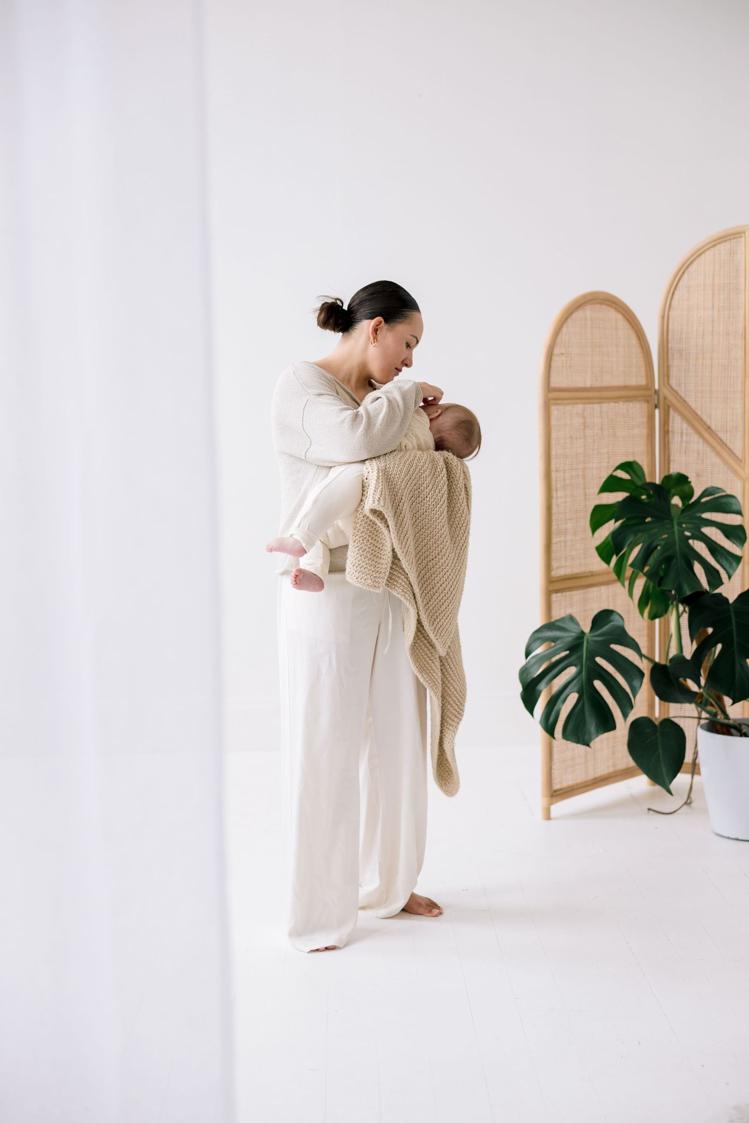 A mother in white outfit tenderly holds her newborn baby wrapped in a beige knit blanket in a bright minimalist room.
