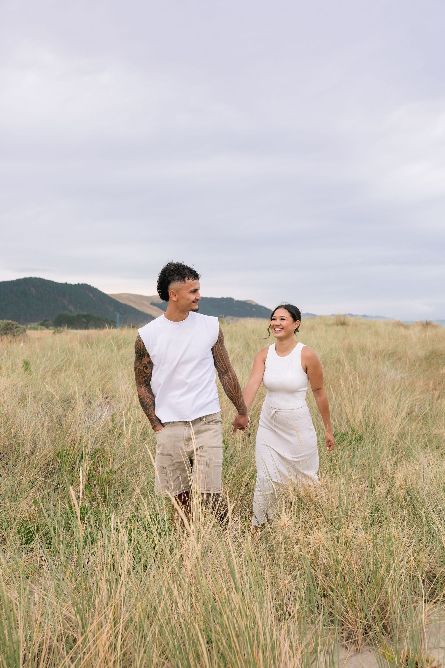 A couple walking hand in hand through a scenic field of tall grass with mountains in the background.