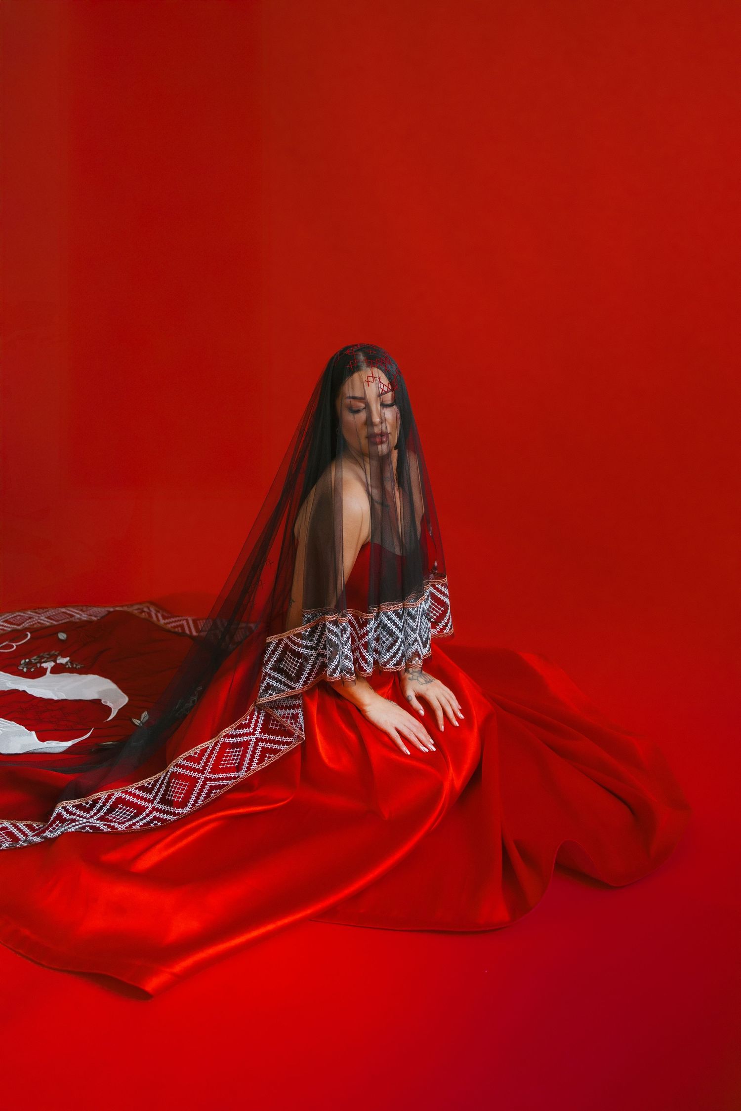 A person in a geometric pattern robe poses on bright red fabric against a red background in an artistic portrait.