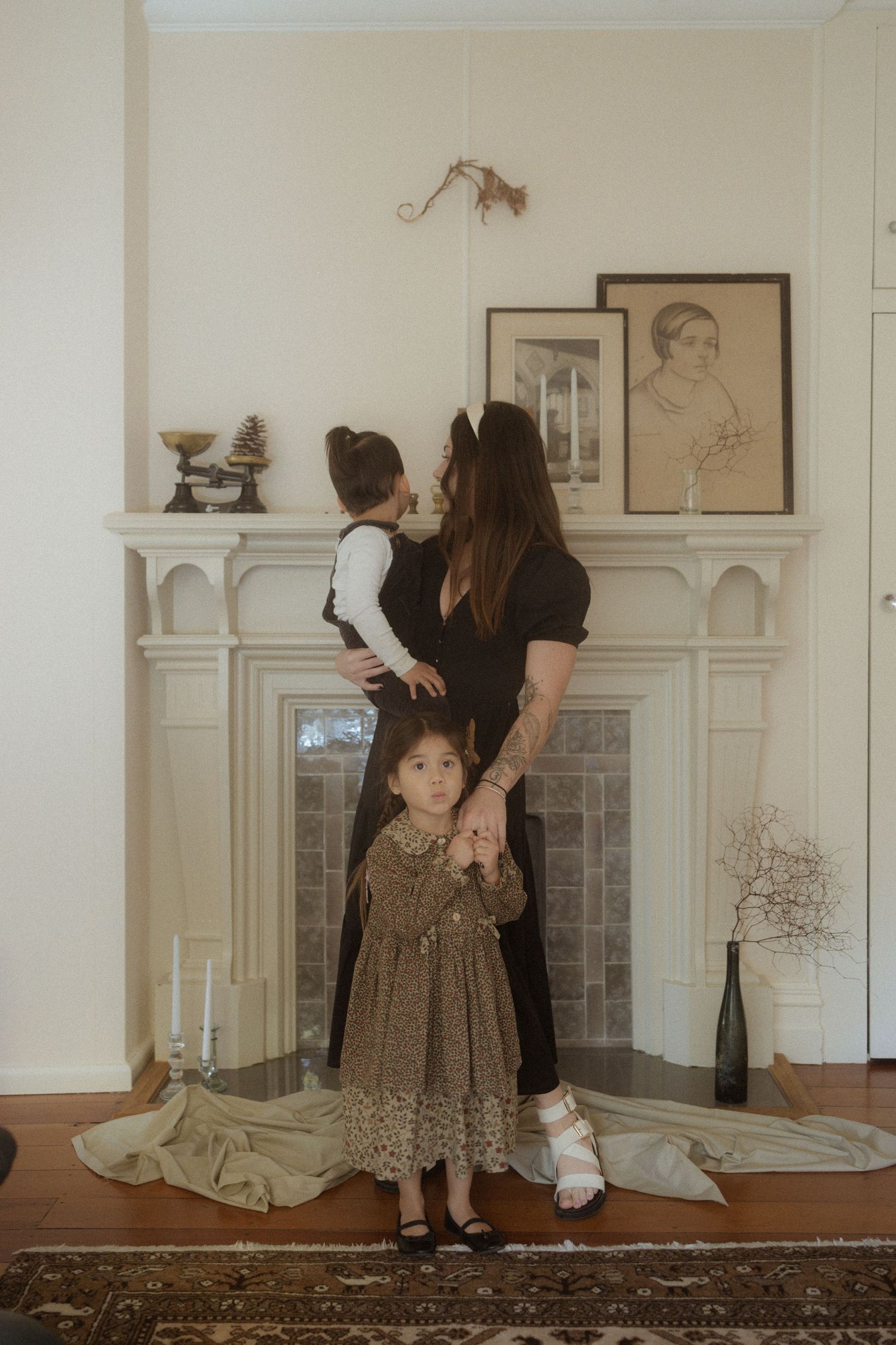 A vintage-styled indoor family scene with figures standing by a decorative fireplace mantel with framed artwork and candles.