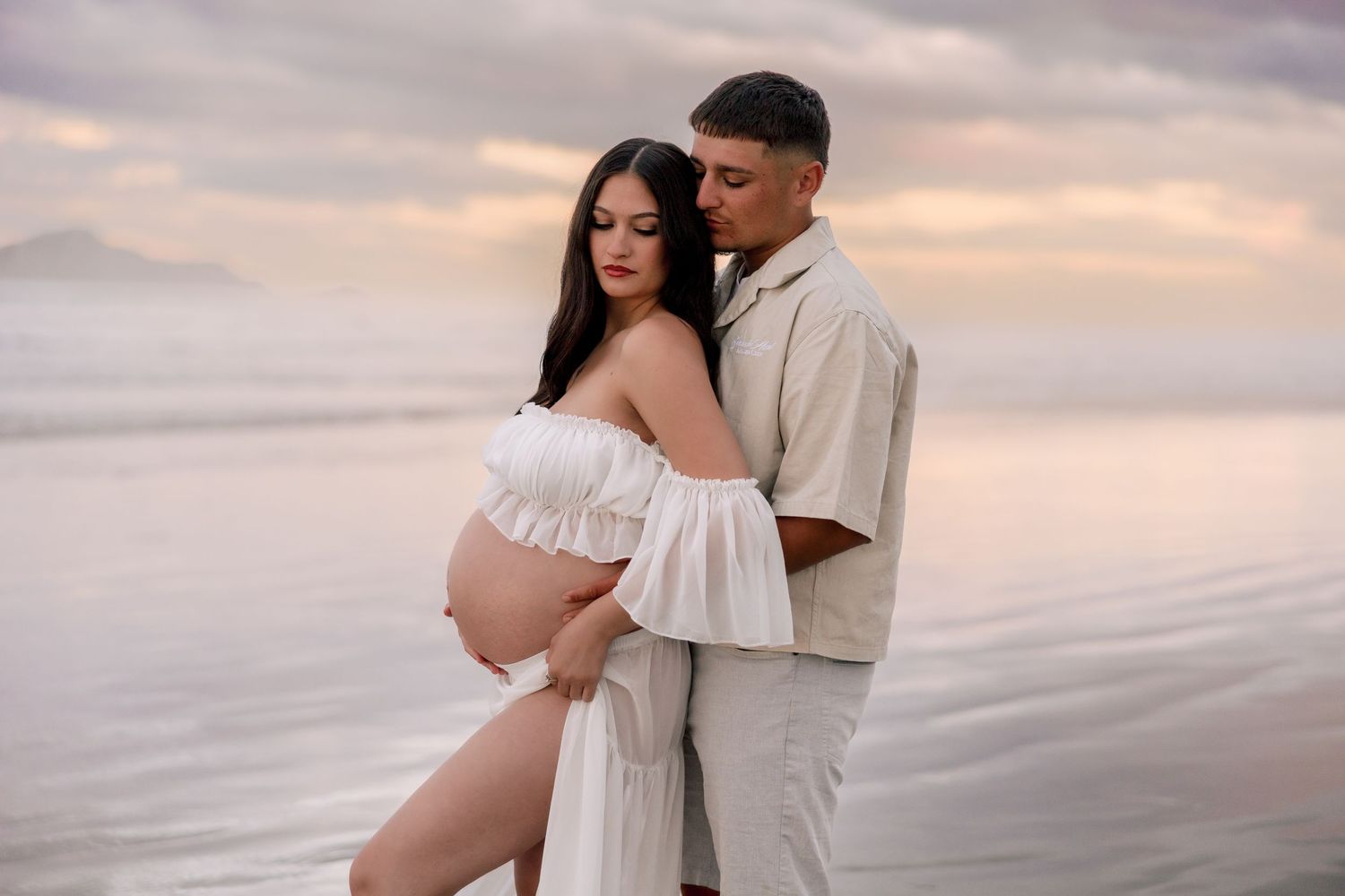 A couple embraces on a serene beach at sunset, with the pregnant woman wearing a flowing white dress.