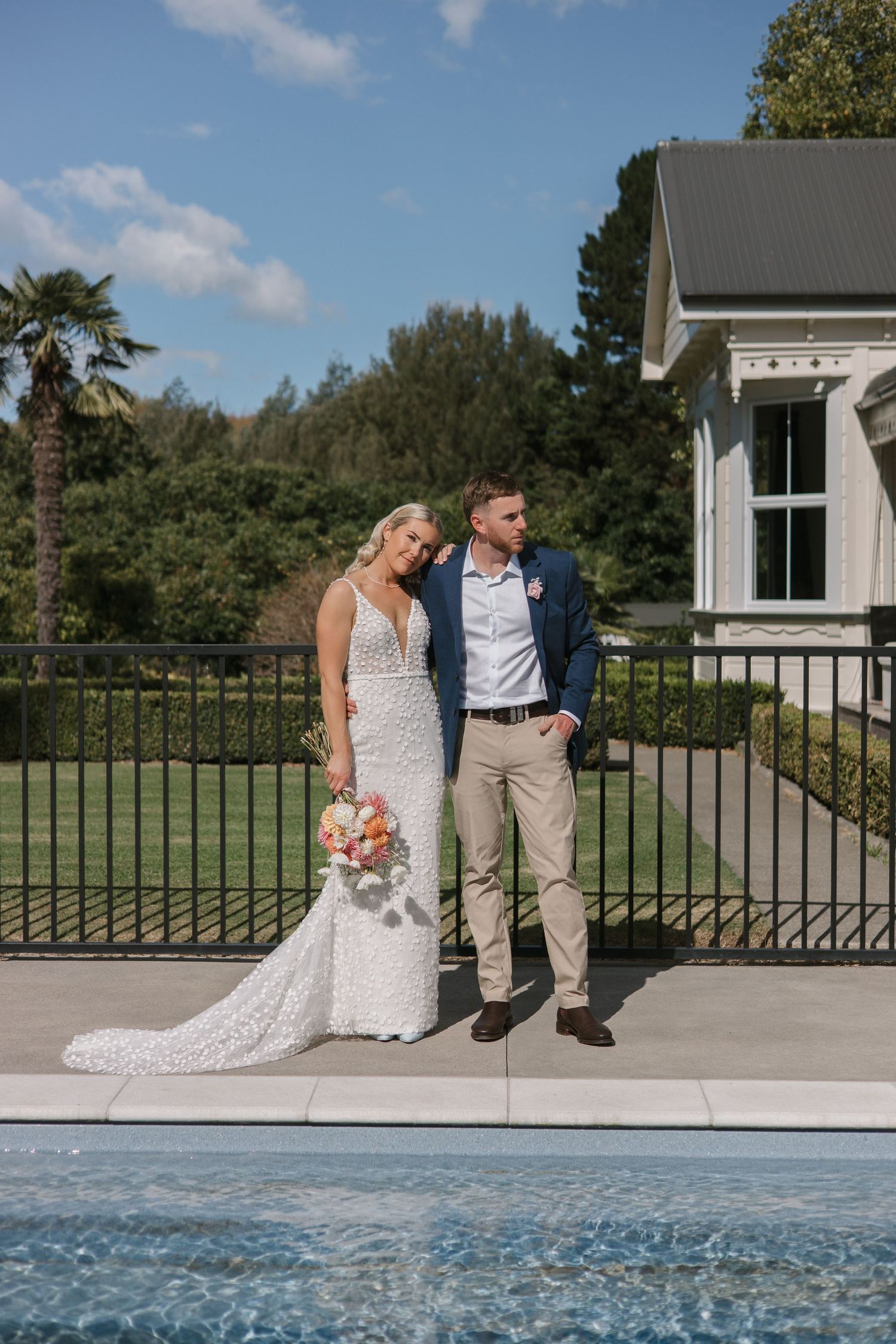 A couple sits together on the edge of a pool deck during a sunny outdoor wedding photoshoot.