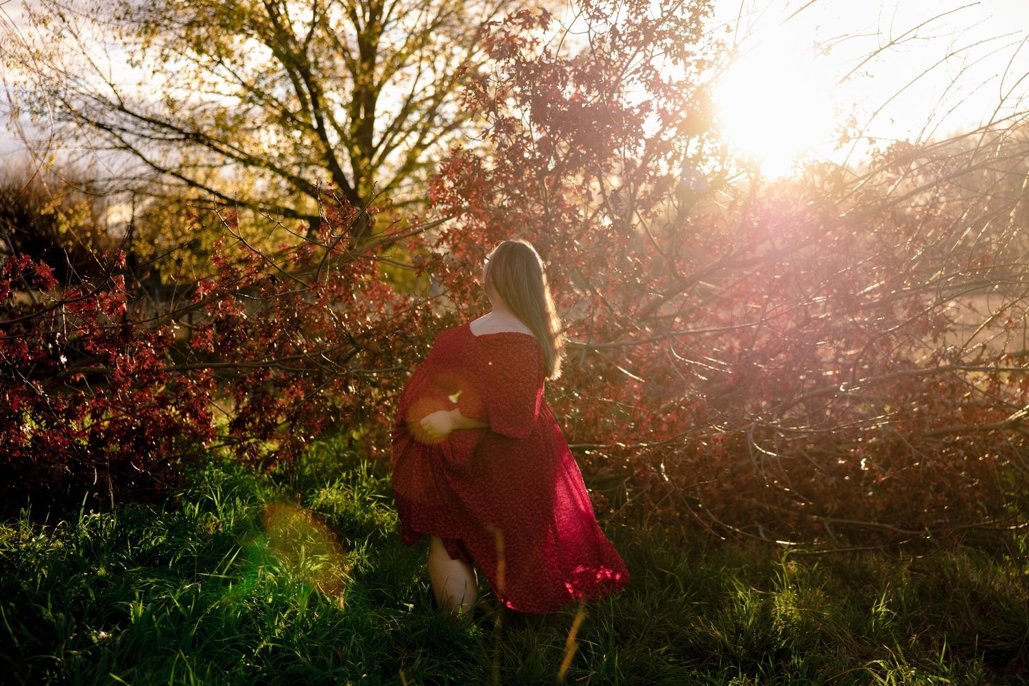 Woman in red dress walking through sunlit autumn forest with golden light filtering through colorful foliage.