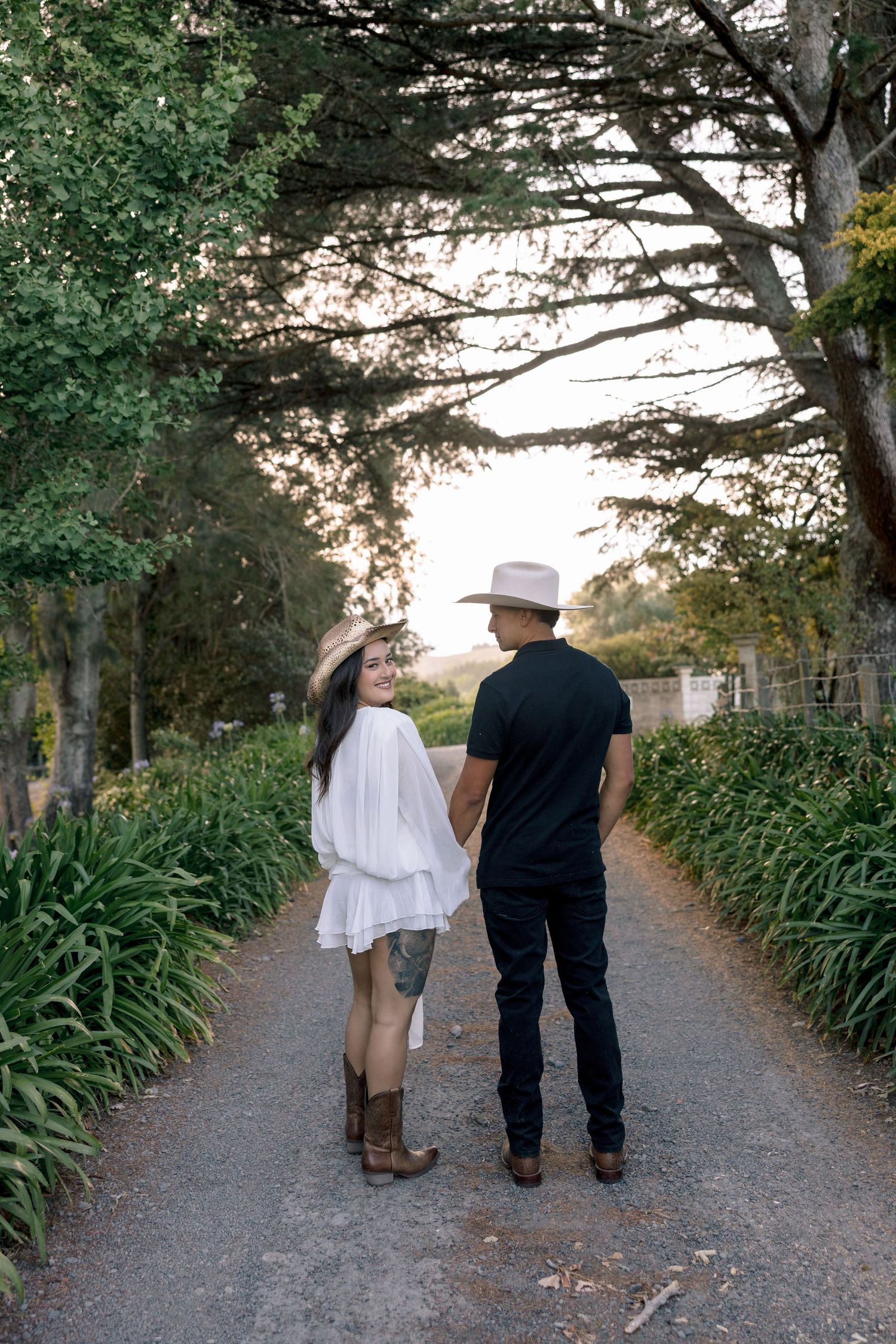 A couple walks down a tree-lined path with lush green foliage on both sides at sunset.