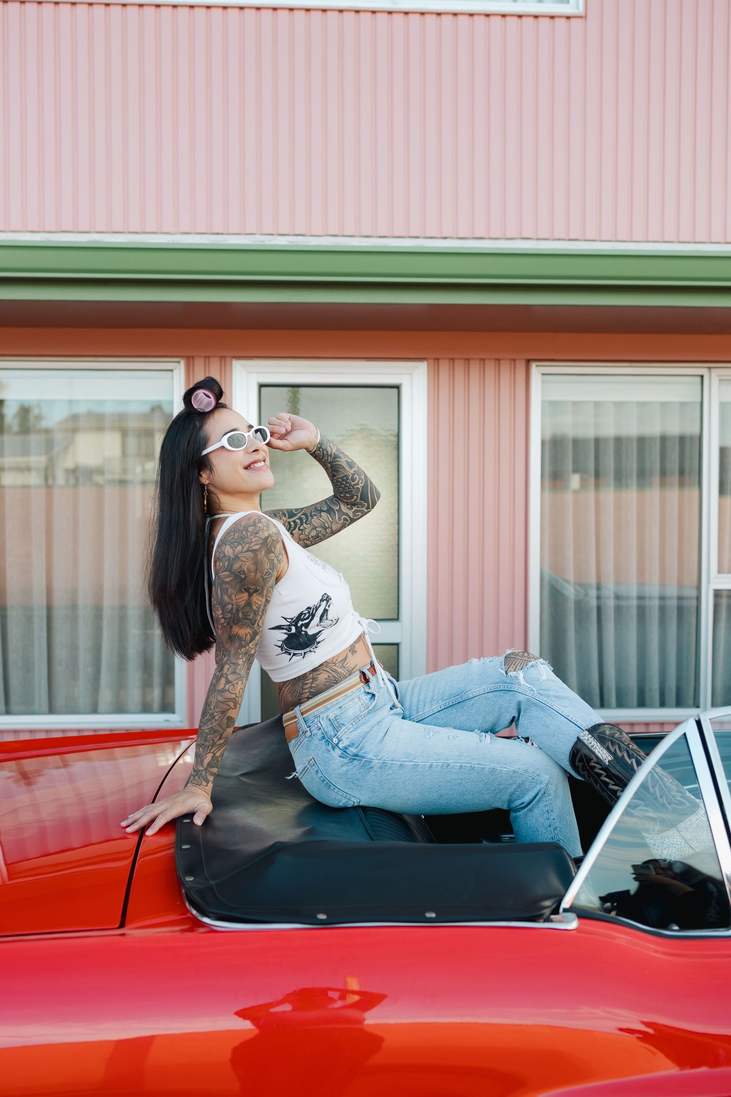 Woman with tattoos sitting on hood of red classic car at retro motel.