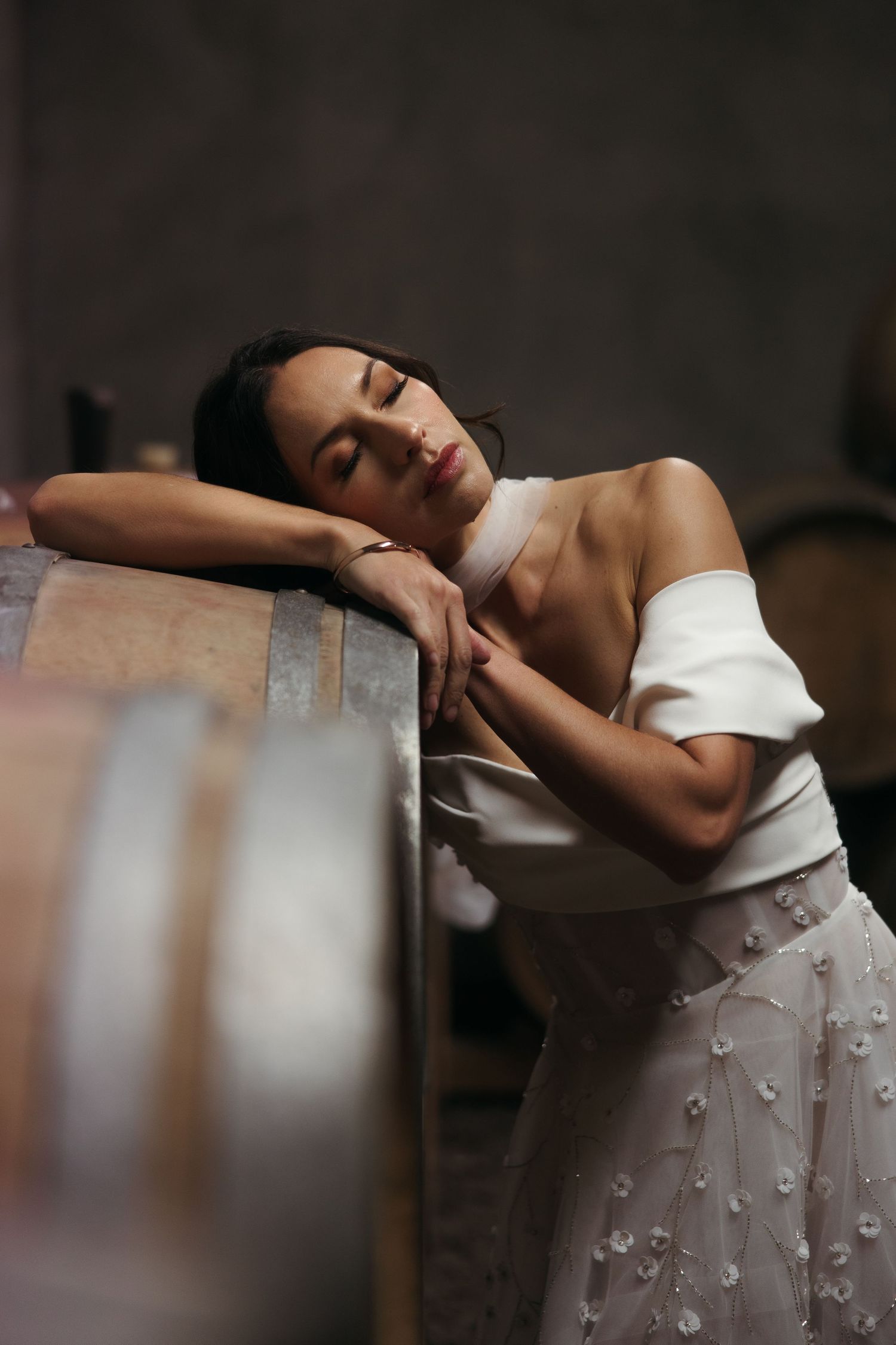 A graceful figure in a white dress with textural details rests dreamily against wooden wine barrels in dim lighting.
