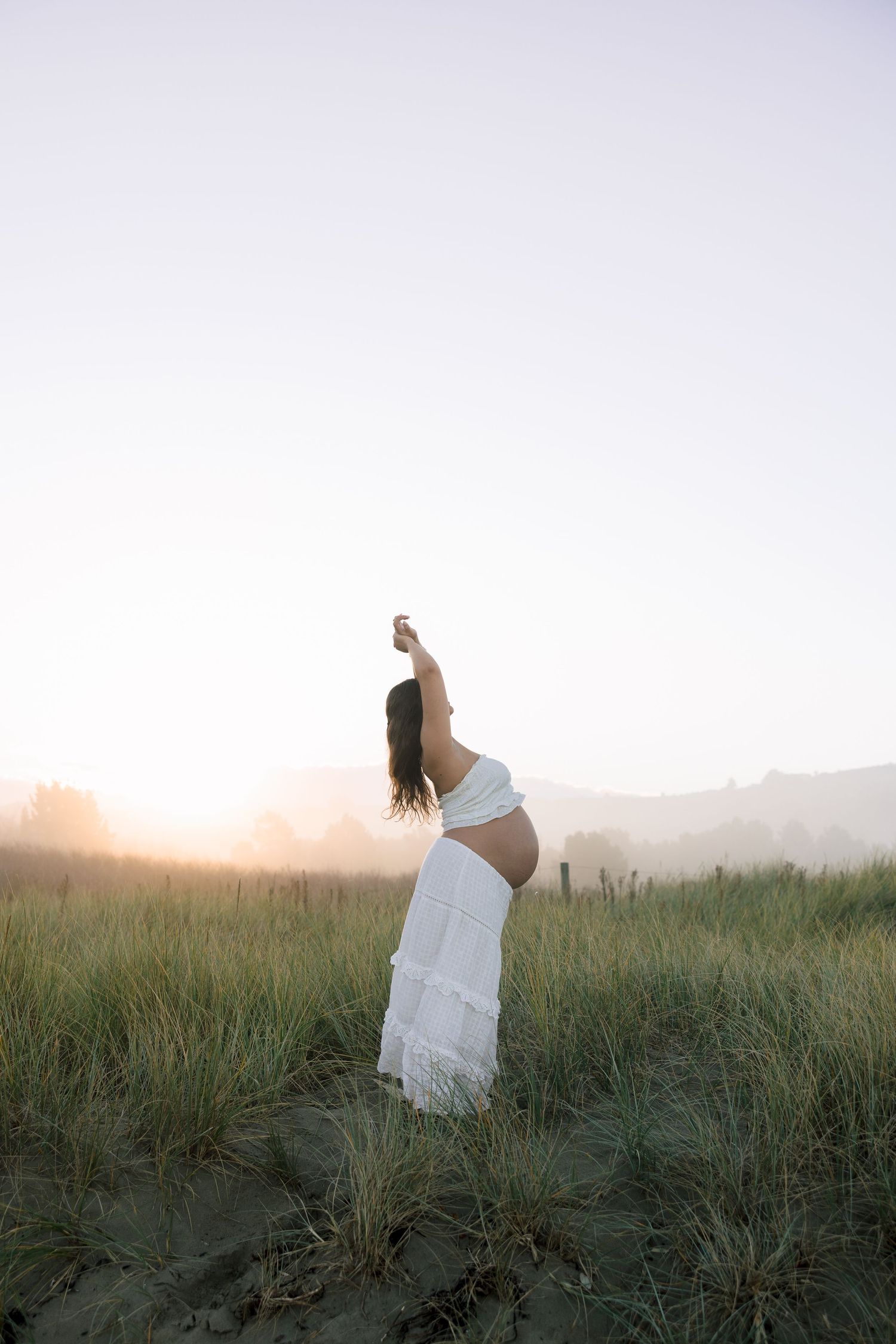 Pregnant woman in white crop top and skirt stands in misty field at golden hour with arm raised.