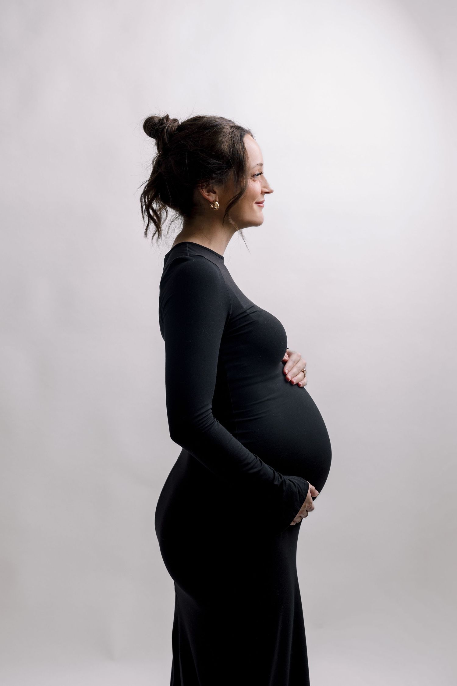 Smiling pregnant woman in black long-sleeve dress cradling baby bump, side profile shot on white background.