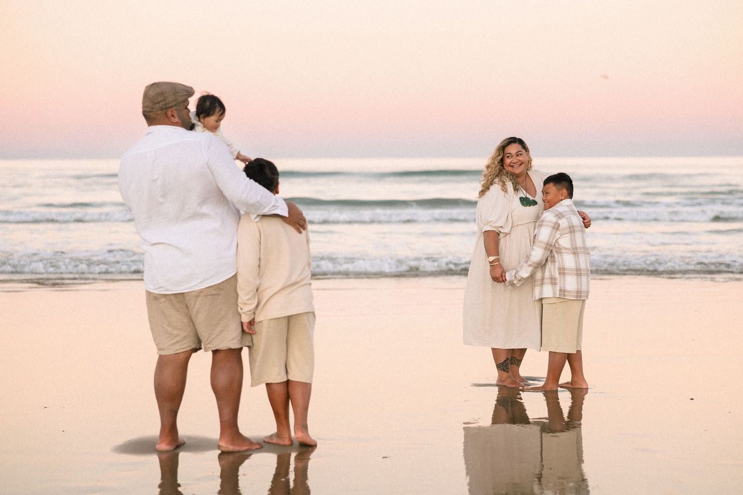 Family in neutral tones poses on a sandy beach at sunset with gentle ocean waves in the background.