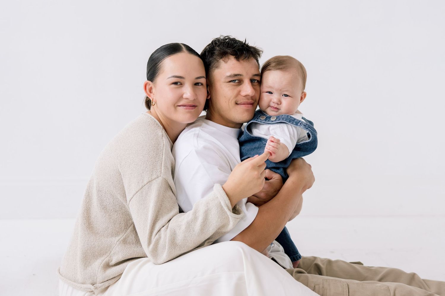 A happy young family with mother, father, and baby boy posing together against a white background.