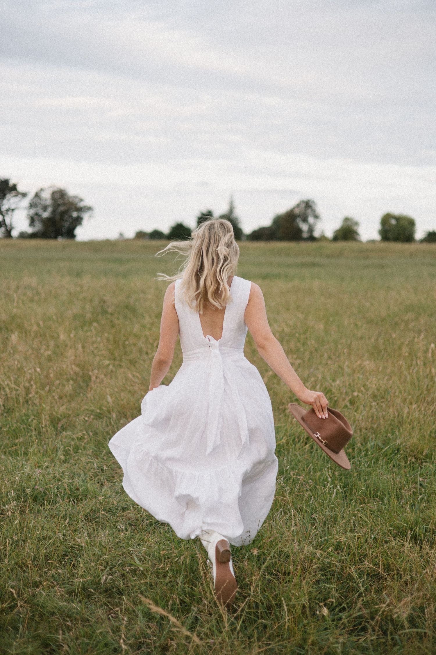 A person in a white dress runs through a sunlit meadow with tall grass on an overcast day.