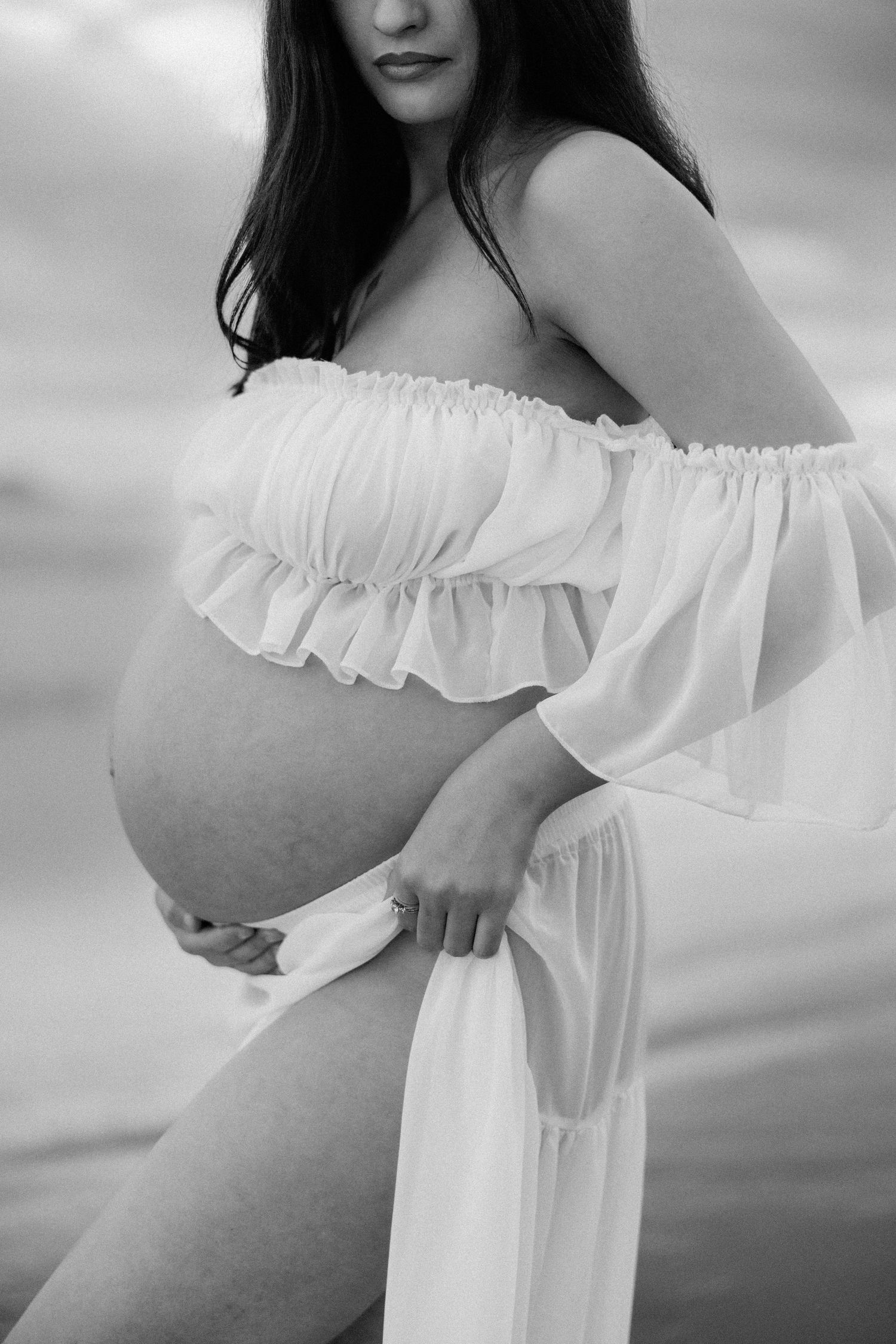 Black and white maternity photo showing a pregnant woman in a flowing white off-shoulder dress against a soft background.