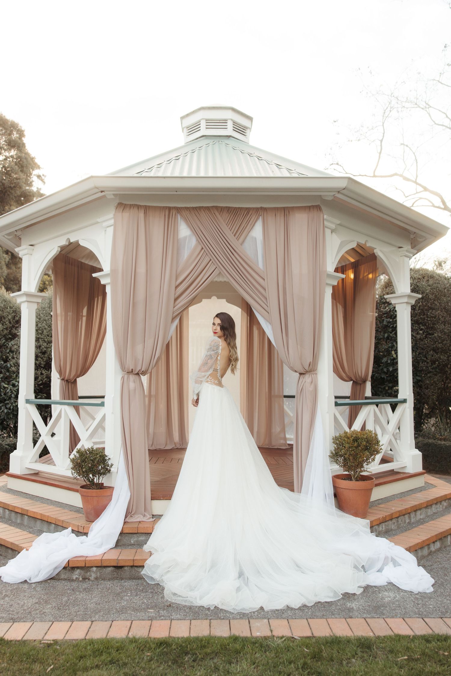 Elegant white bridal gown flowing around a figure posed in a garden gazebo draped with blush chiffon curtains.