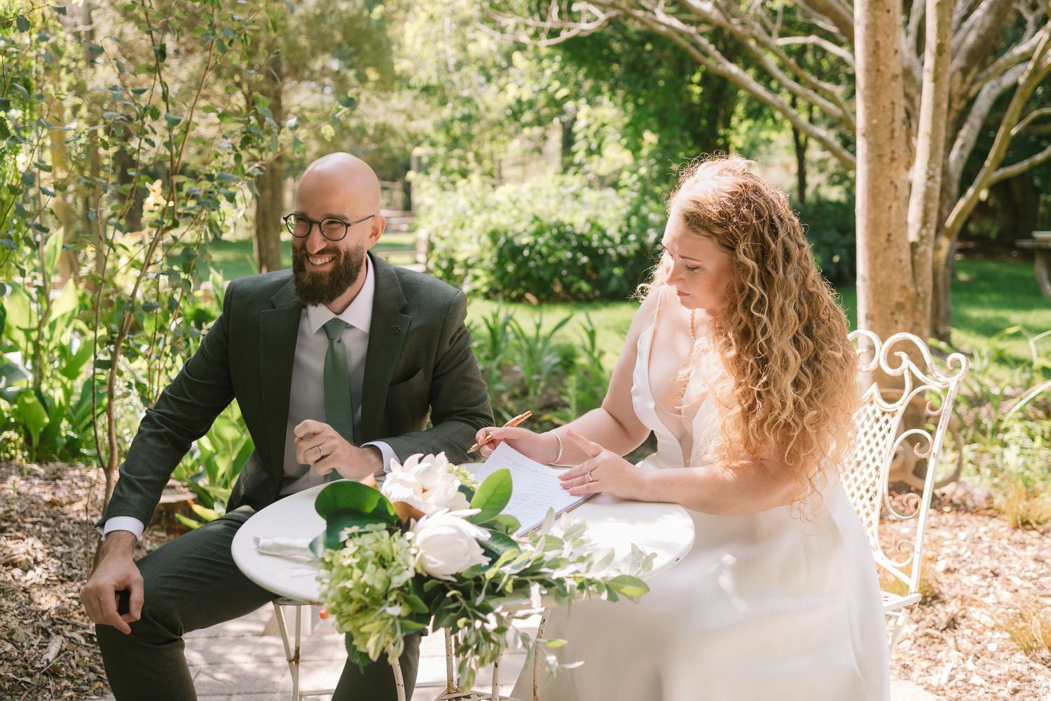 Newlyweds sitting at outdoor garden table with white floral centerpiece.