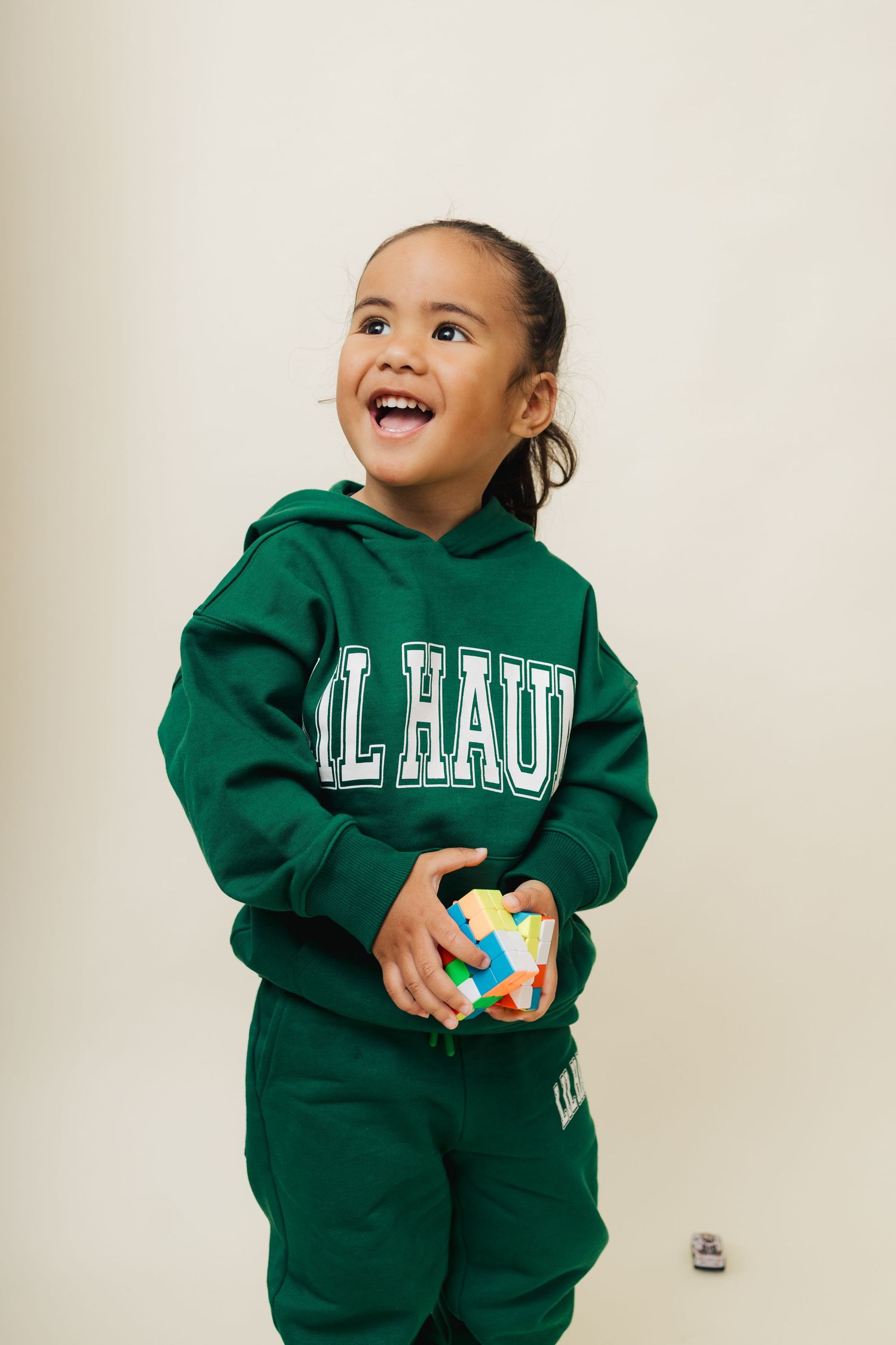 Child wearing a green al hau sweatsuit holds a colorful puzzle cube while looking up with a joyful expression.