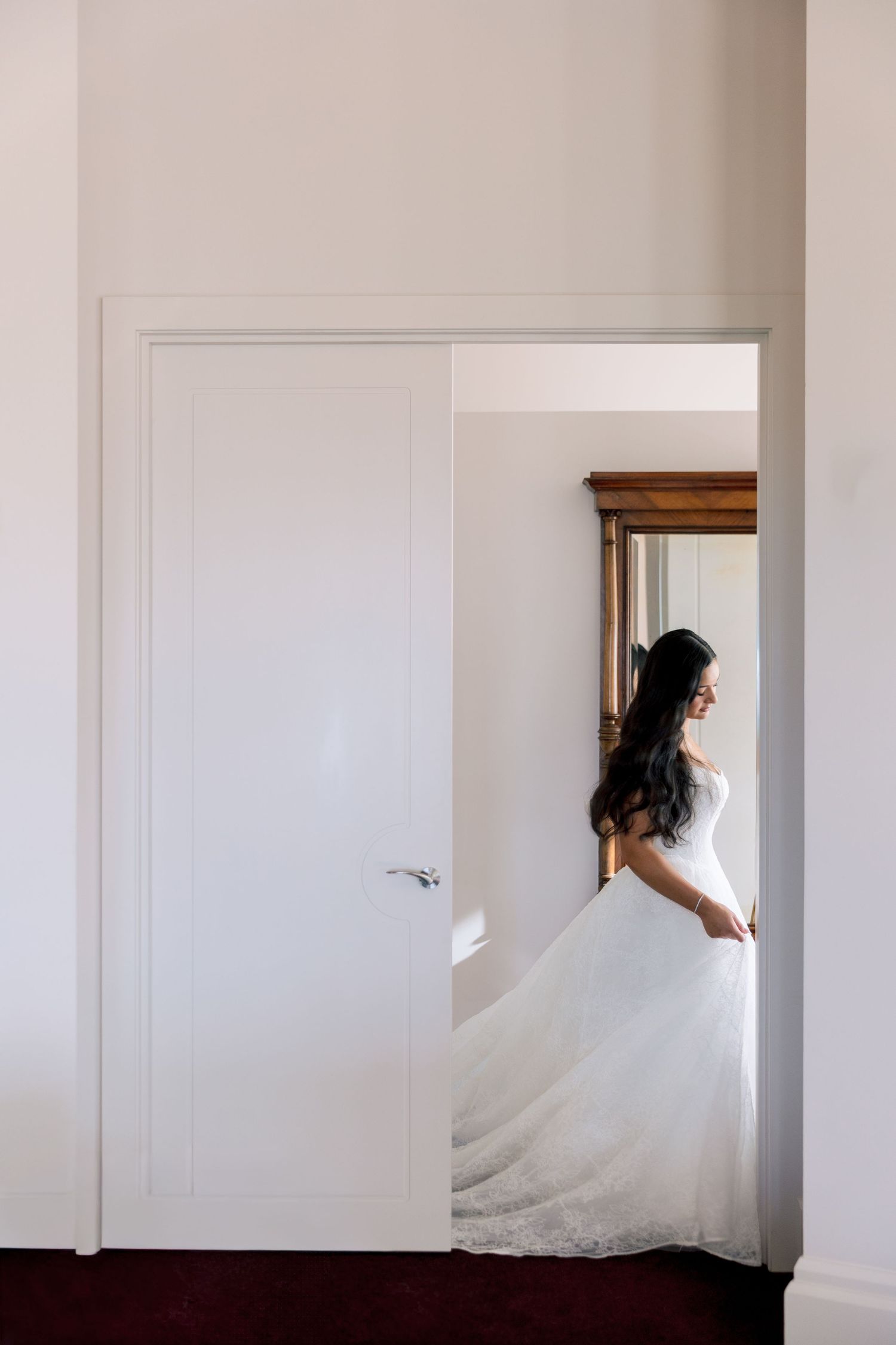 Bride in elegant white wedding gown with long dark hair standing near a white door in a bright room.
