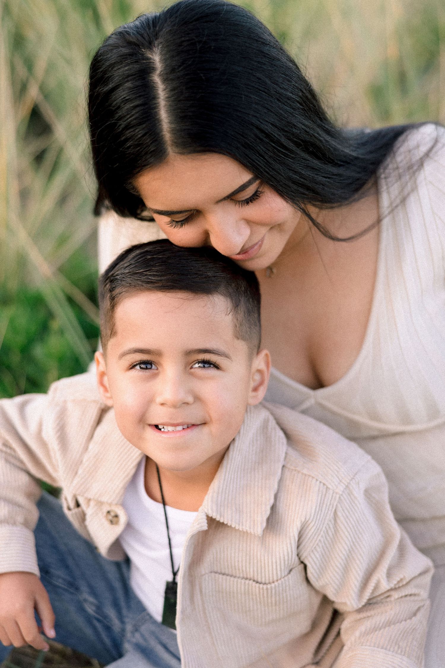 A loving mother leans over her smiling young son wearing a beige shirt outdoors.