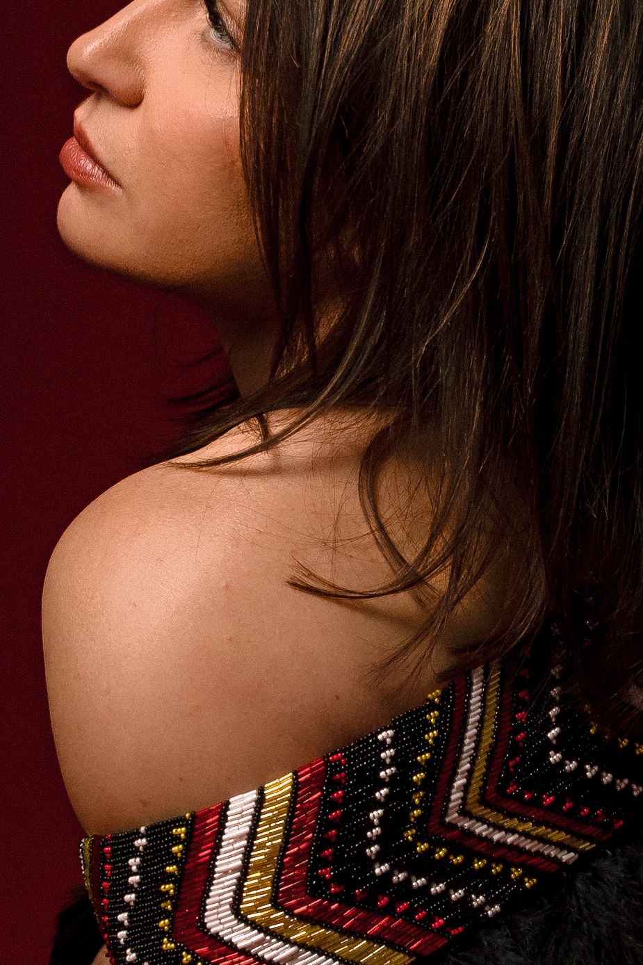 Young woman in profile view wearing a colorful beaded off-shoulder top against a dark red background.
