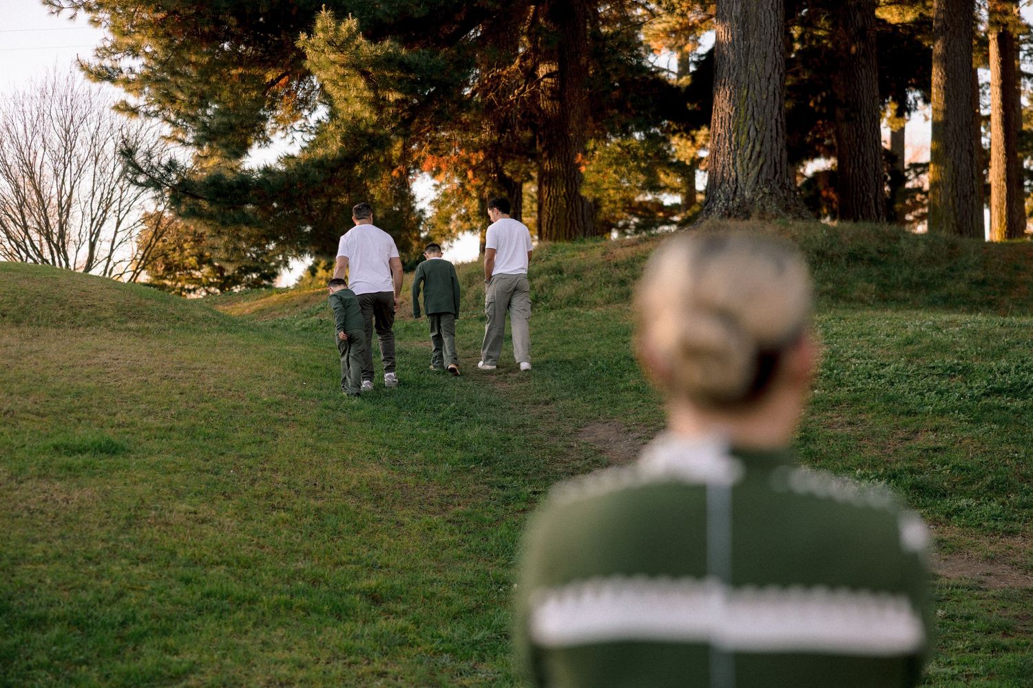 People walking up a grassy hill toward tall trees in a park at golden hour, viewed from behind a person in a green jacket.