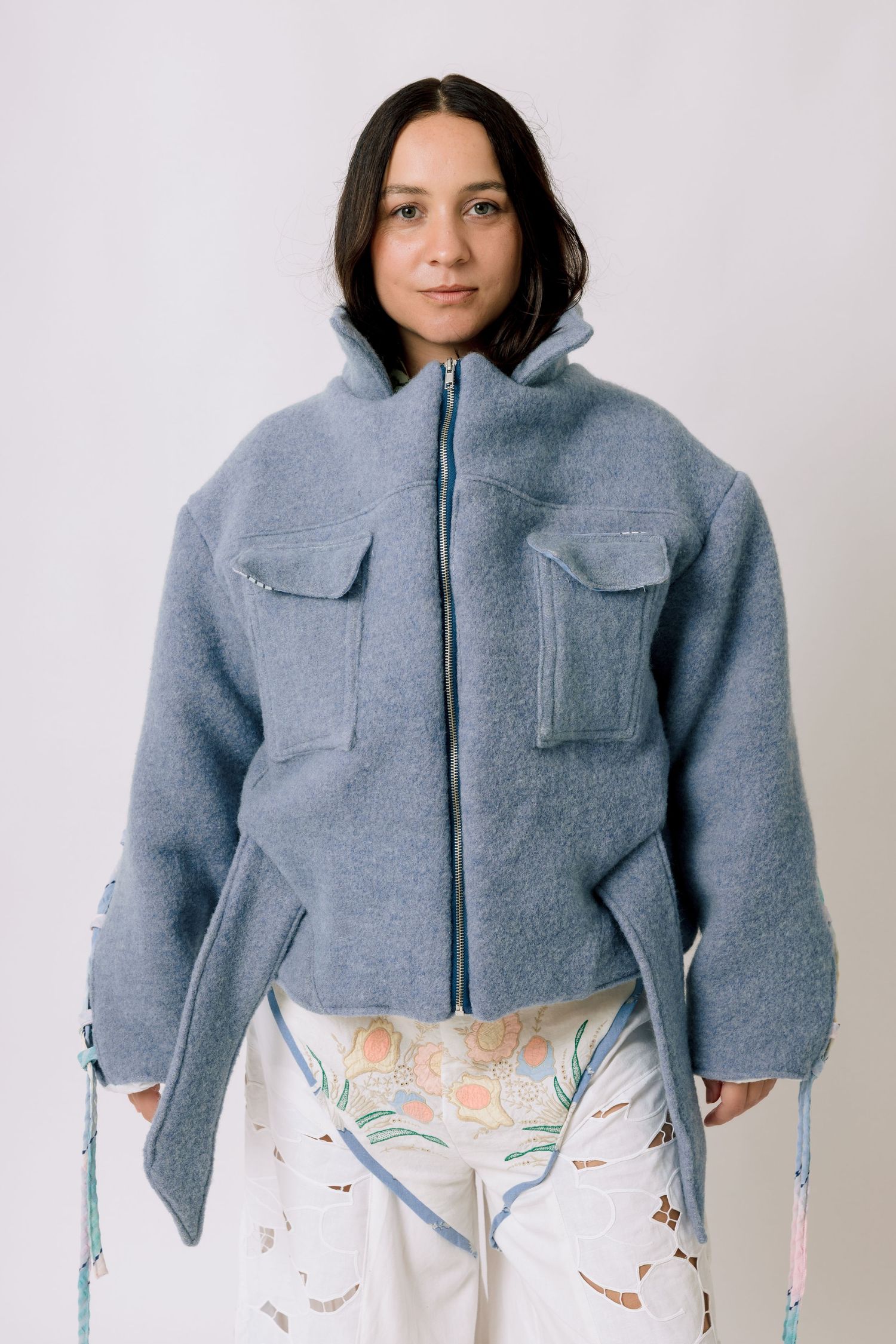 A person wearing a gray wool zip-up jacket with utility pockets and white patterned pants against a white background.