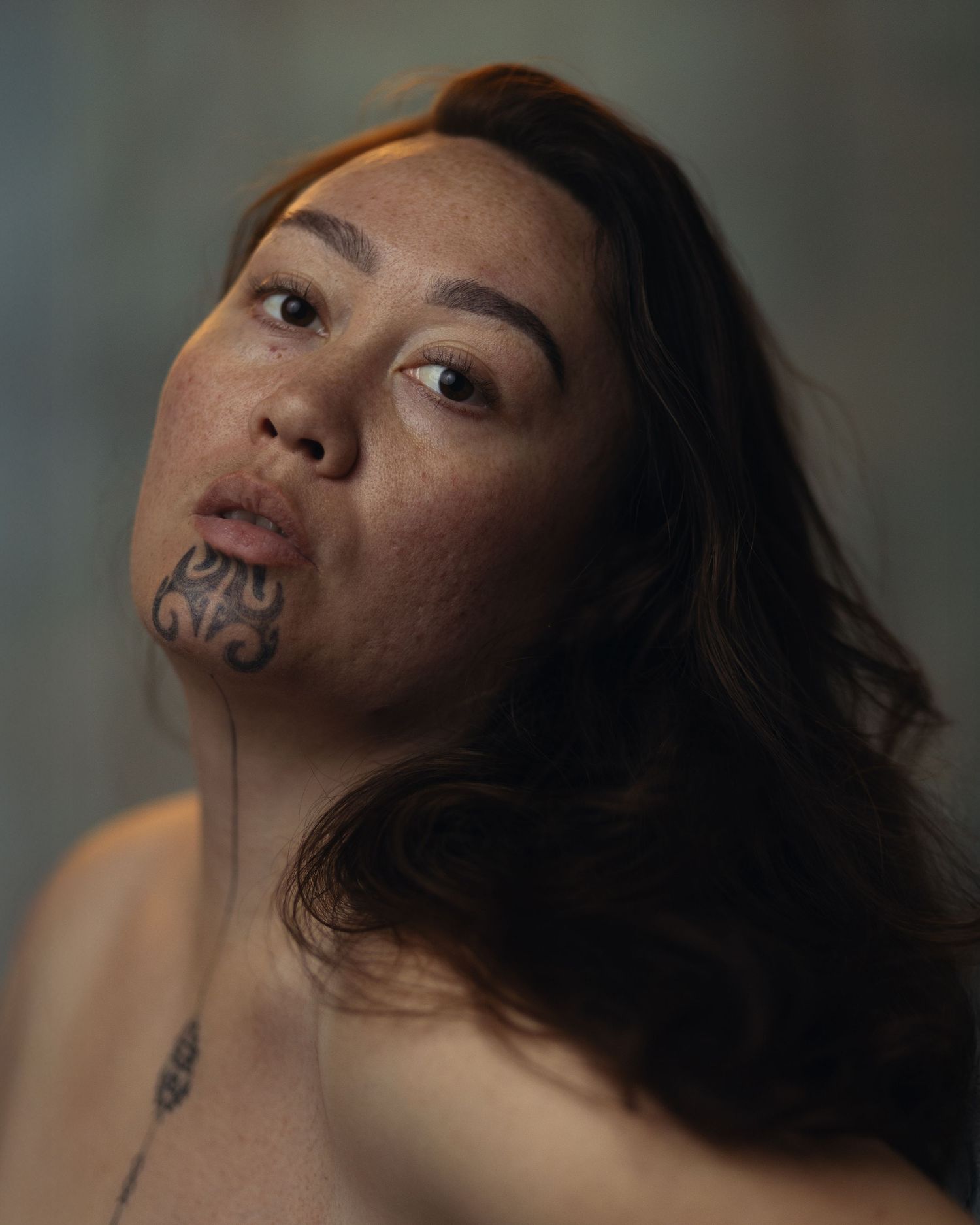 Portrait of a woman with traditional Maori ta moko chin tattoo and long dark hair against neutral background.