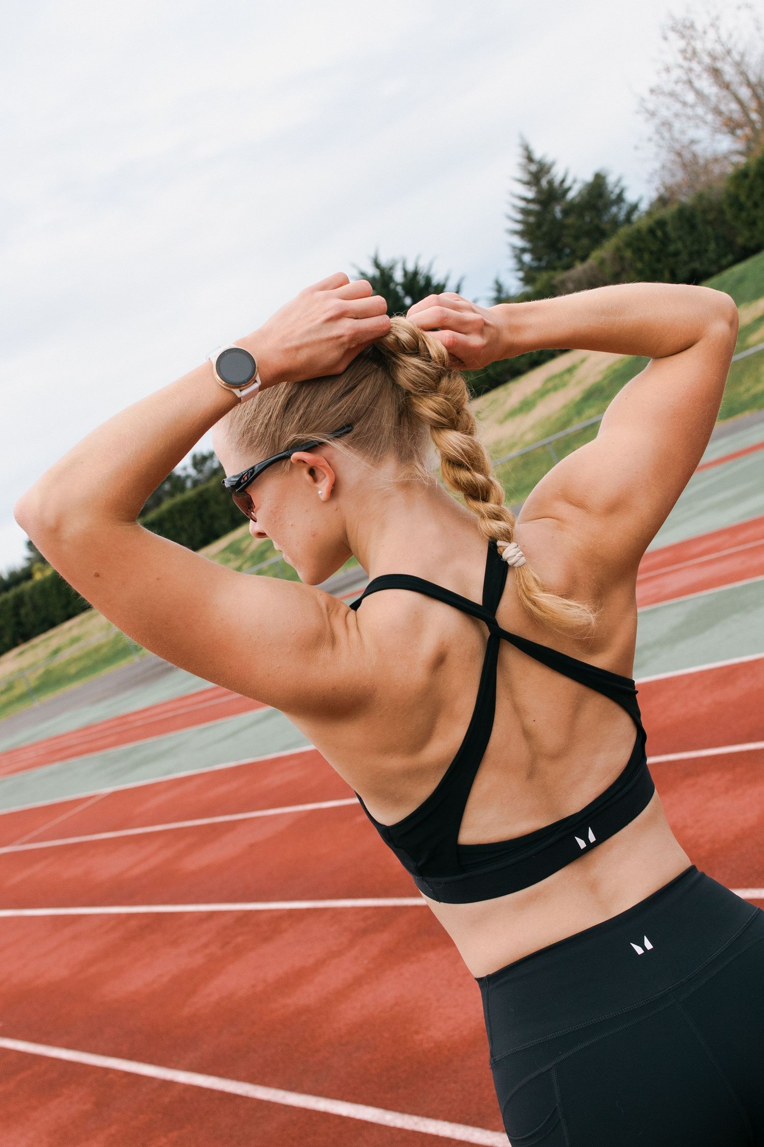 A female athlete stretches her arms and back at an outdoor running track in black athletic wear.