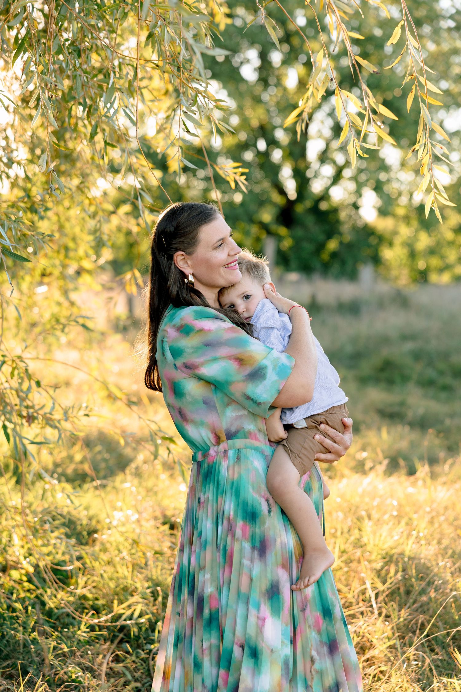 Mother in colorful dress hugging smiling toddler outdoors in golden sunlight near willow tree.