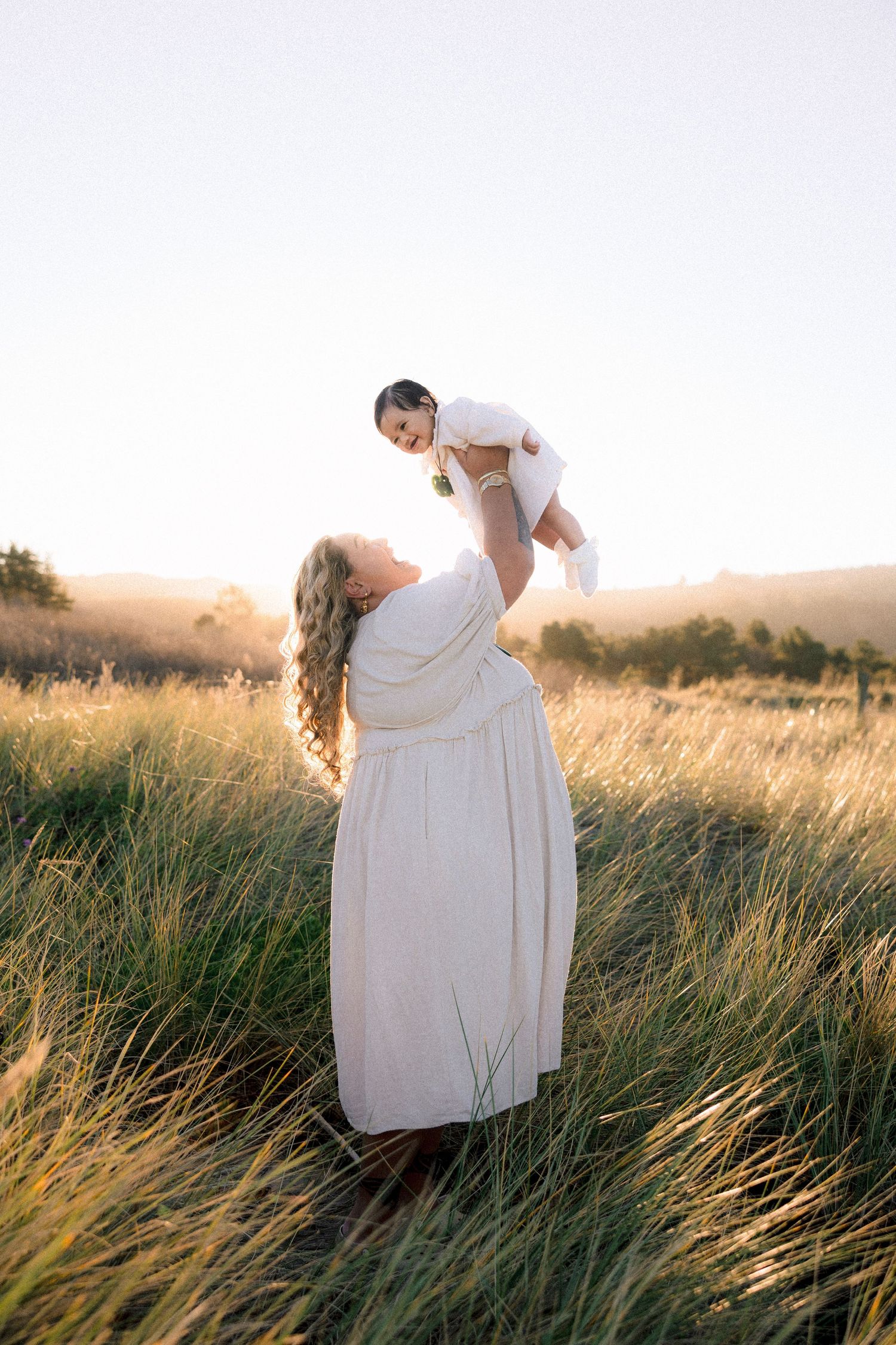 Mother in white dress lifting baby in sunlit golden field at sunset.