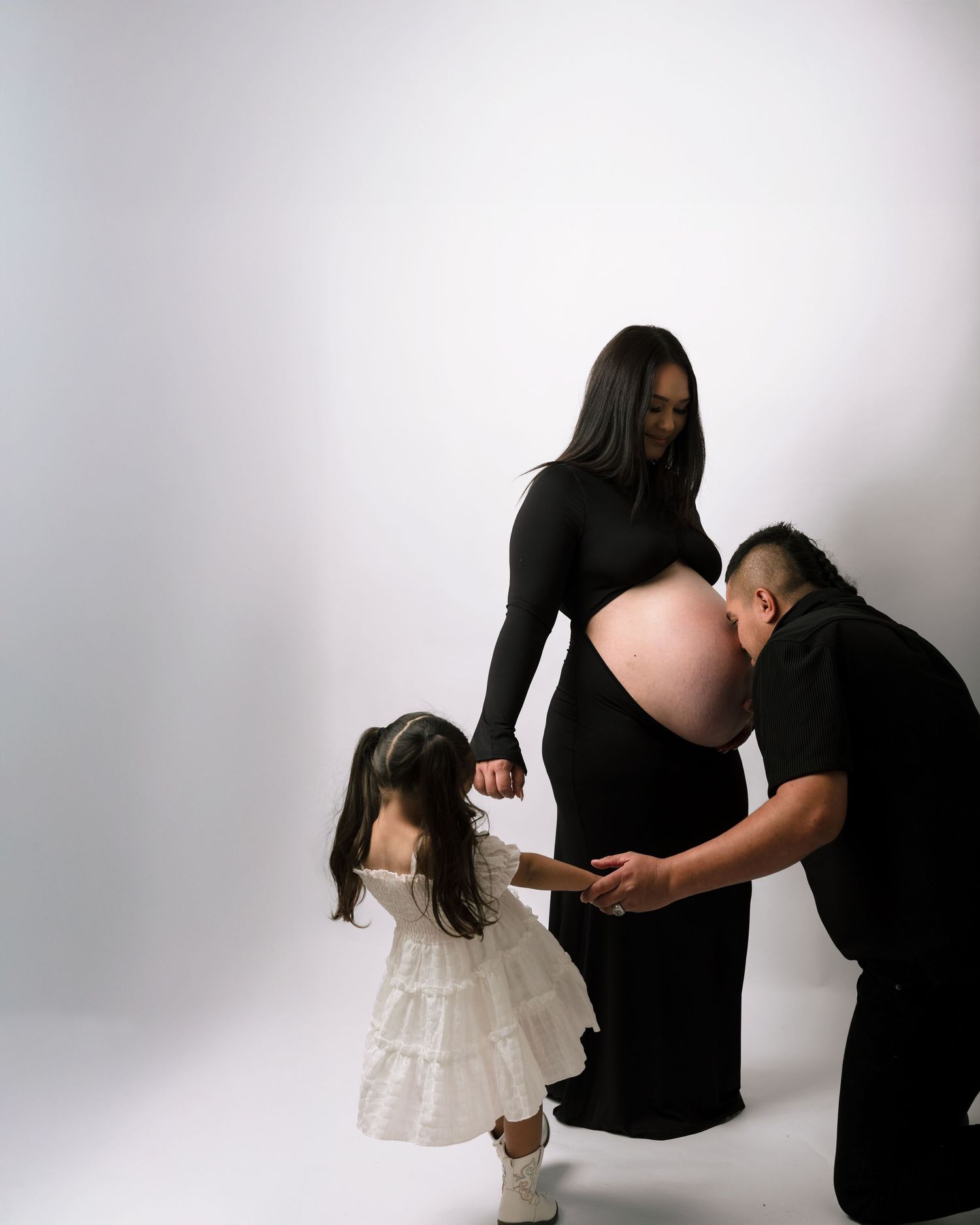 Pregnant woman in black dress poses with young daughter in white dress and partner touching baby bump.