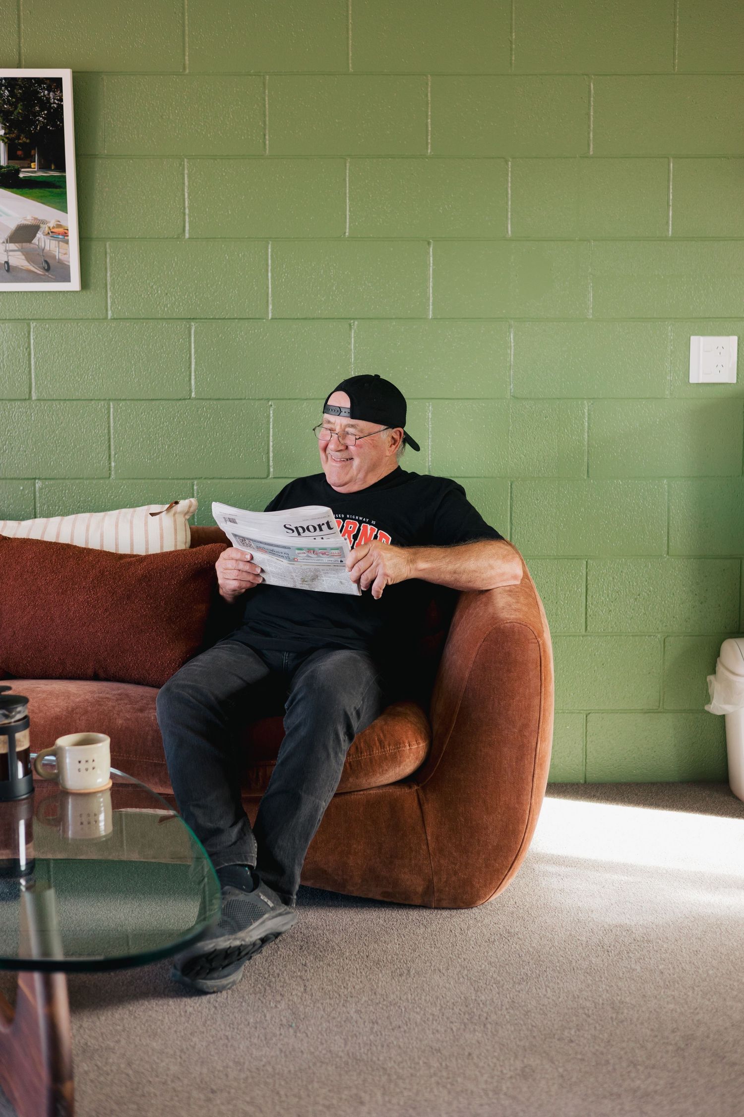 Person relaxing on brown velvet couch reading newspaper against green wall.