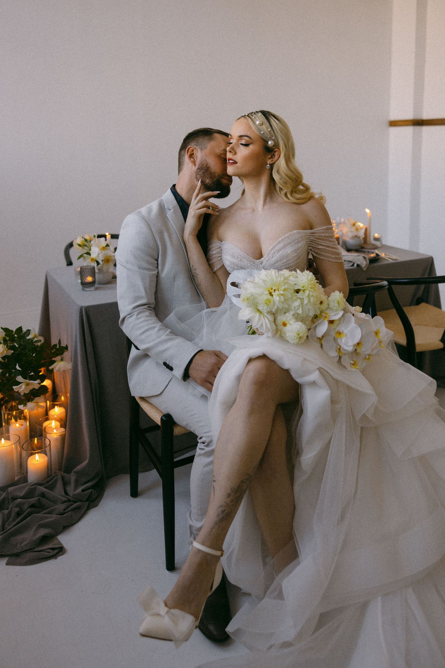 Romantic wedding couple sharing intimate moment on bench surrounded by white flowers and glowing candles.