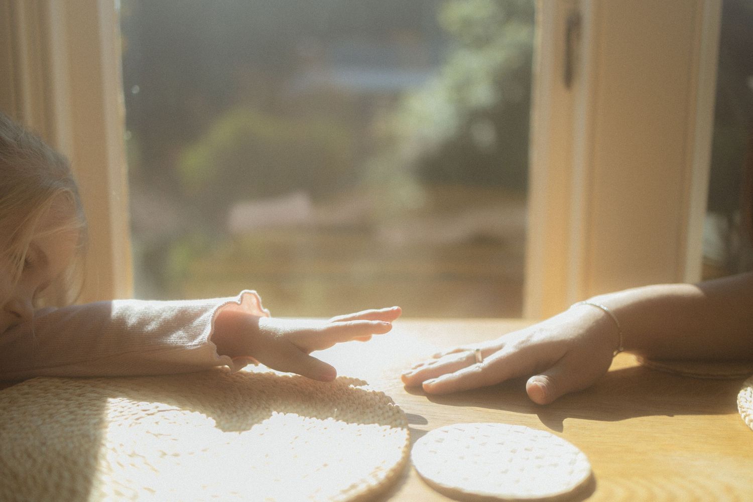 Two hands reaching across a sunlit table with warm rays streaming through a window as breakfast dishes rest between them.