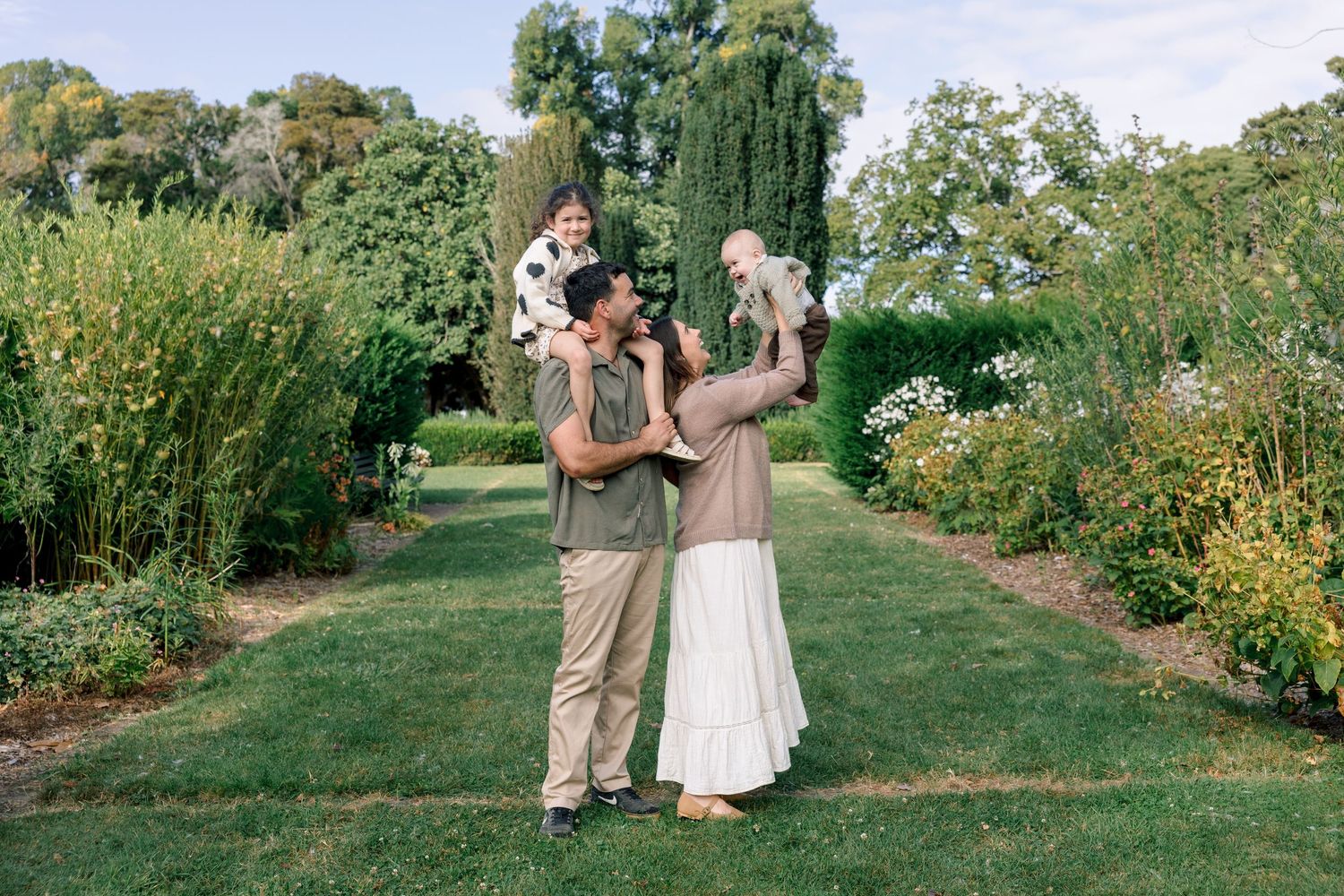 Happy family of four posing in a lush garden path, parents lifting two young children outdoors on a sunny day.