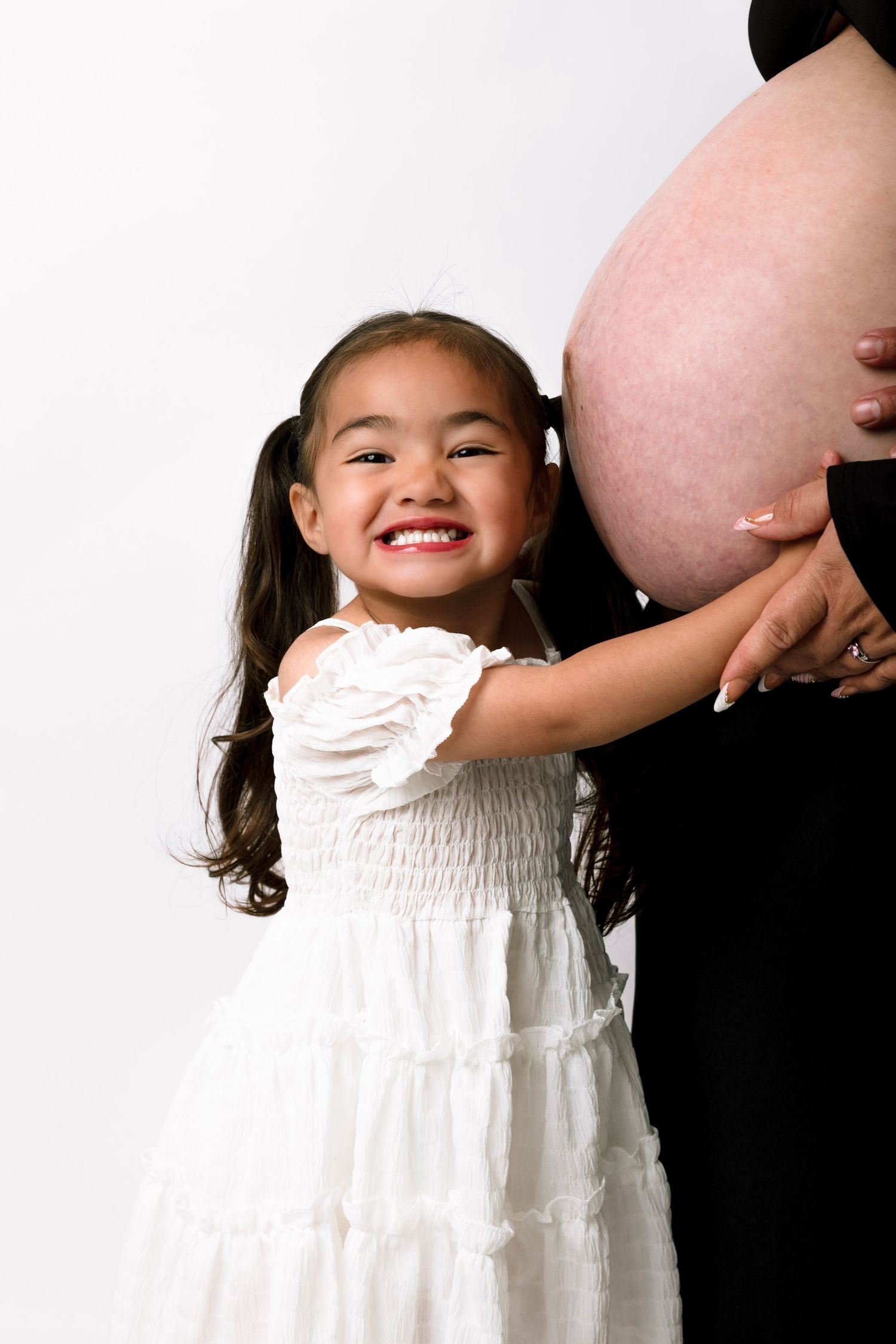 A smiling young girl in a white dress touching her pregnant mother's bare baby bump in a studio photo.