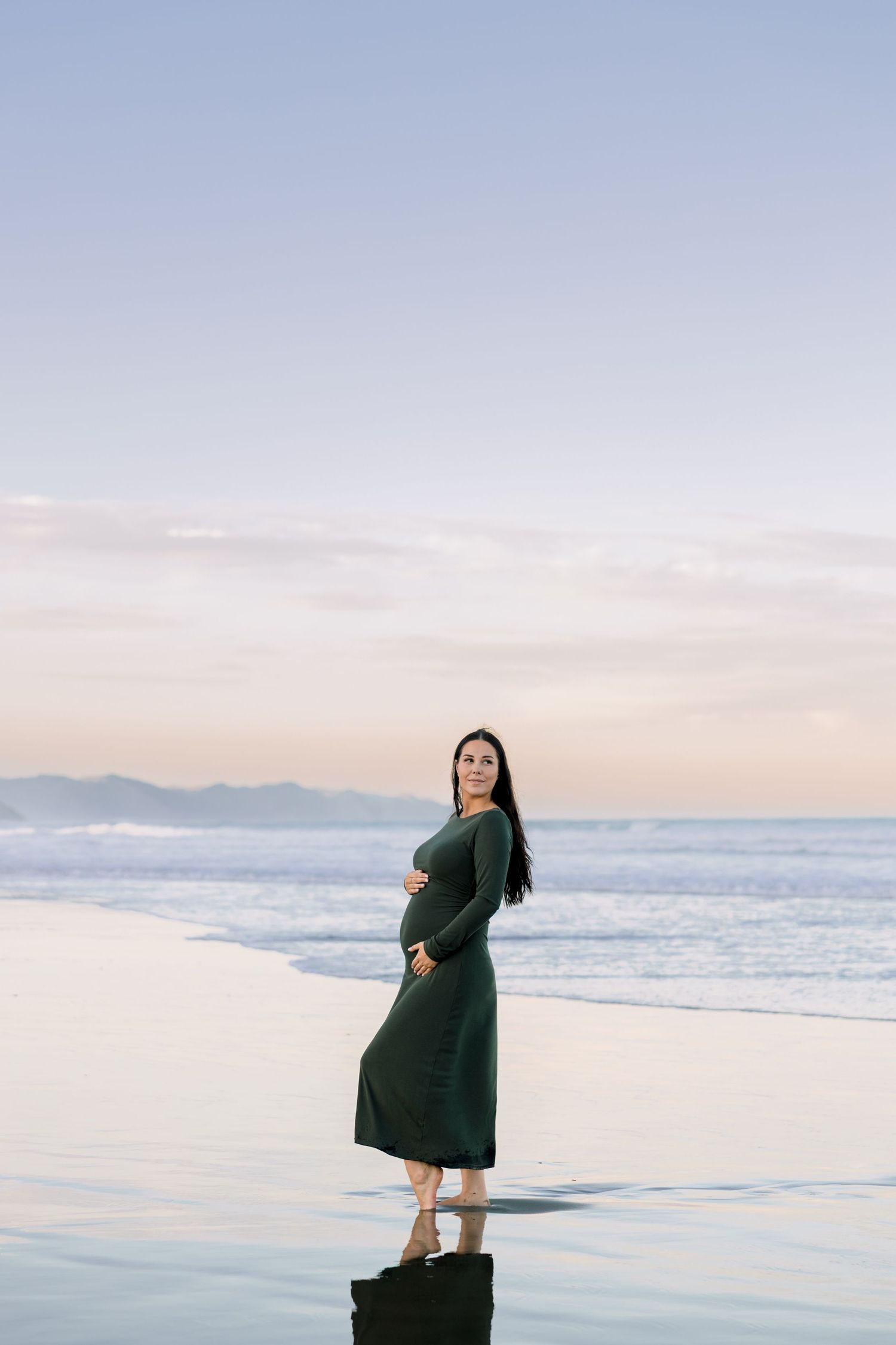 Pregnant woman in green dress standing barefoot on beach at sunset, cradling baby bump by ocean waves.