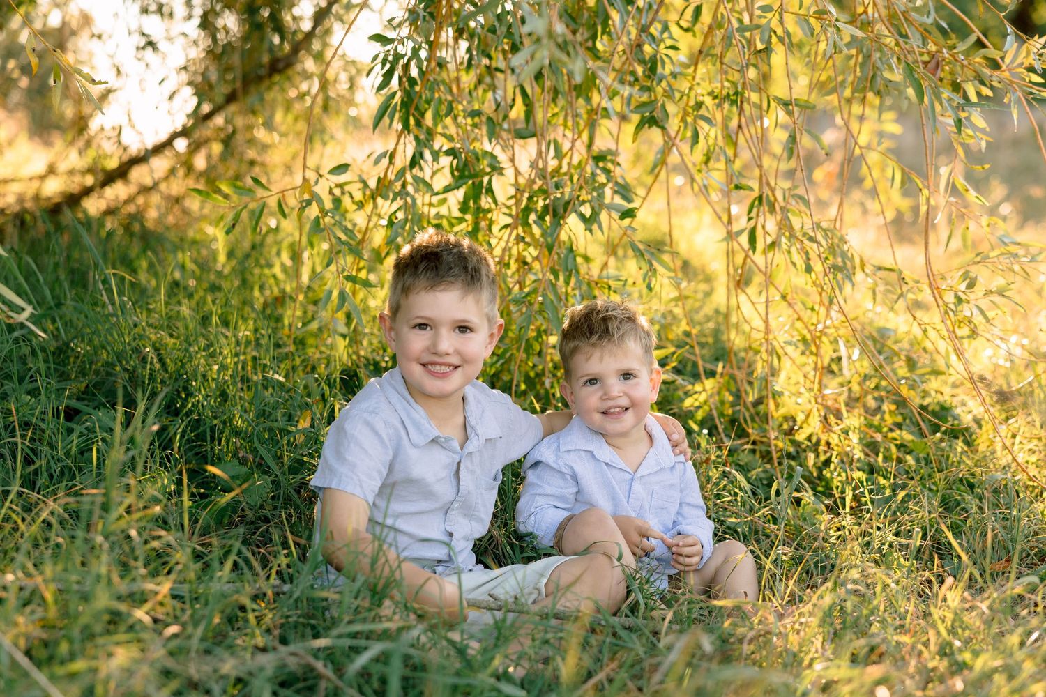 Two smiling brothers in white shirts sitting together in lush green grass under a willow tree in golden sunlight.
