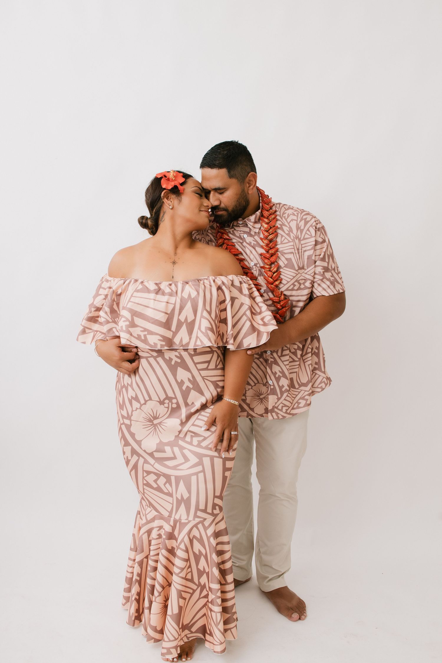 A romantic couple in matching pink Hawaiian style outfits embrace against a white background during a photo session.
