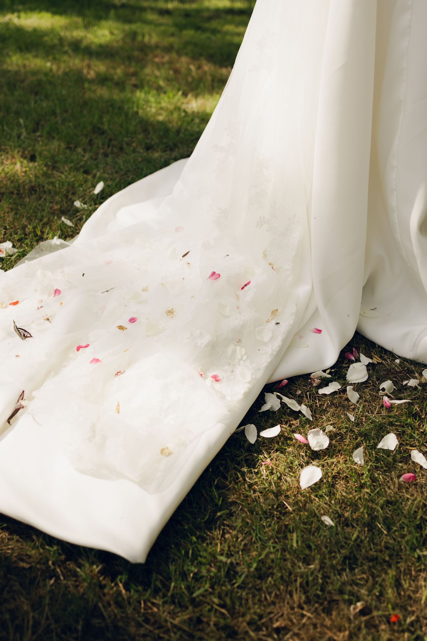 White wedding dress train lying on grass with scattered rose petals.