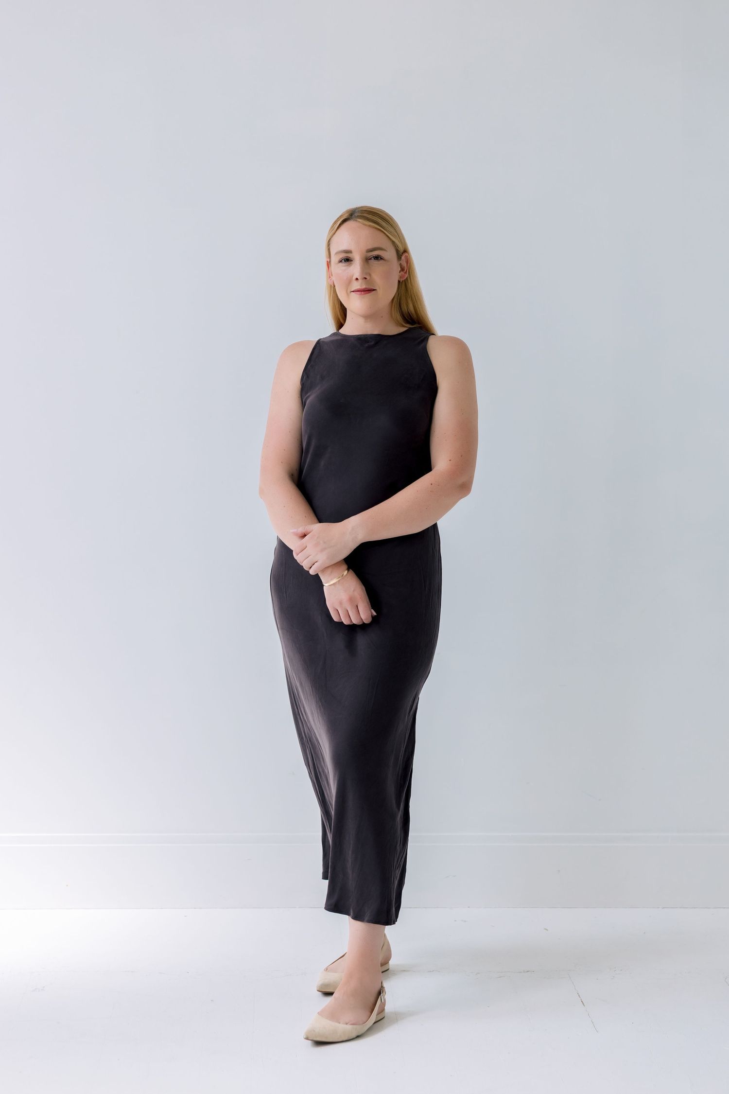Fashion model poses in a black sleeveless dress against a light background in a minimalist studio setting.