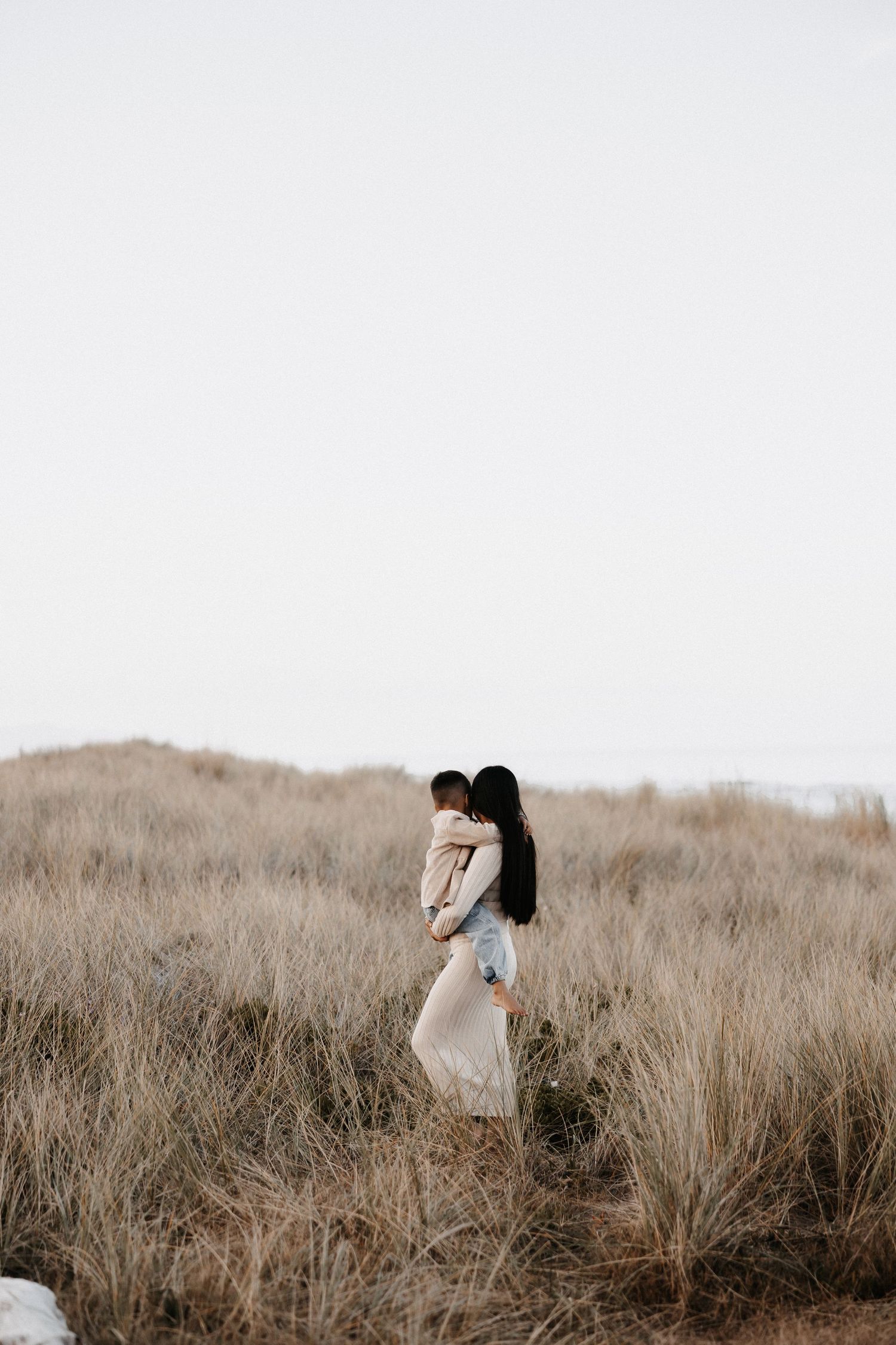A woman in a beige dress holds a child in a golden grass field under a soft, overcast sky.