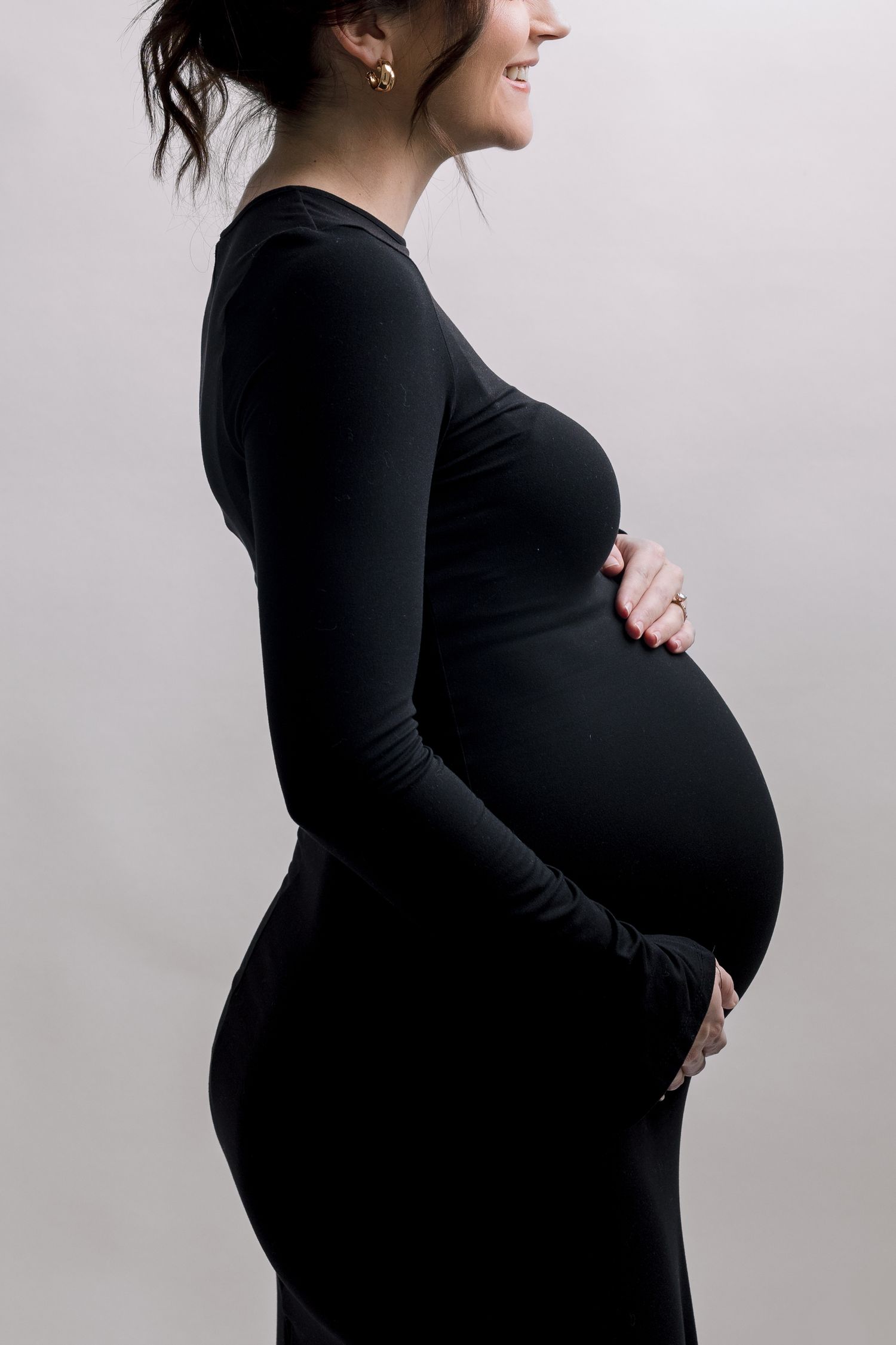 Smiling pregnant woman in black long-sleeve dress cradling baby bump, side profile maternity photo on gray background.