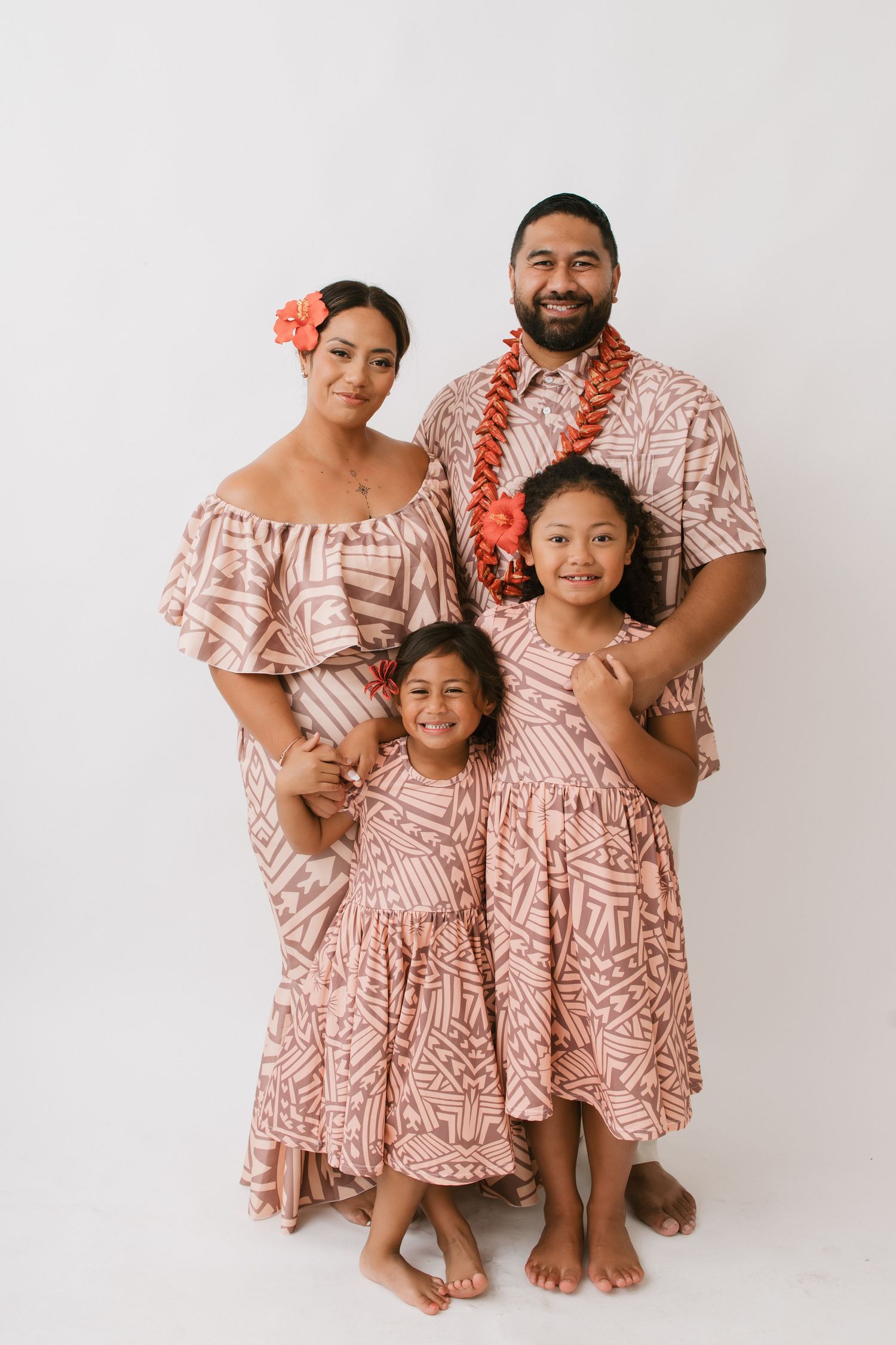 A family wearing matching pink and white patterned Hawaiian outfits poses together barefoot against a white background.