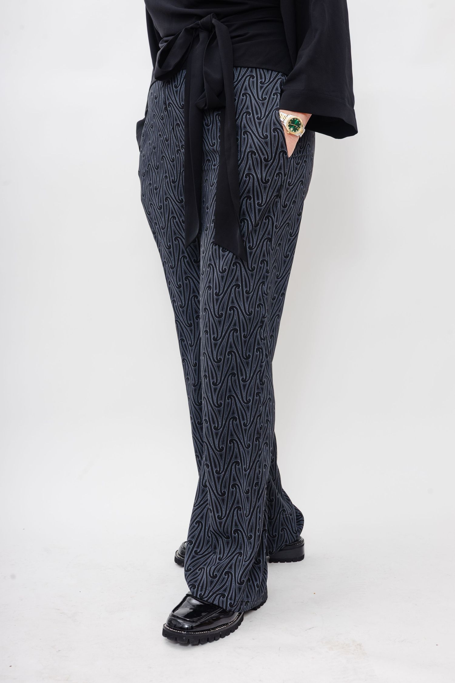A person wearing black and gray patterned wide-leg pants with a tie waist and black shoes against a white background.